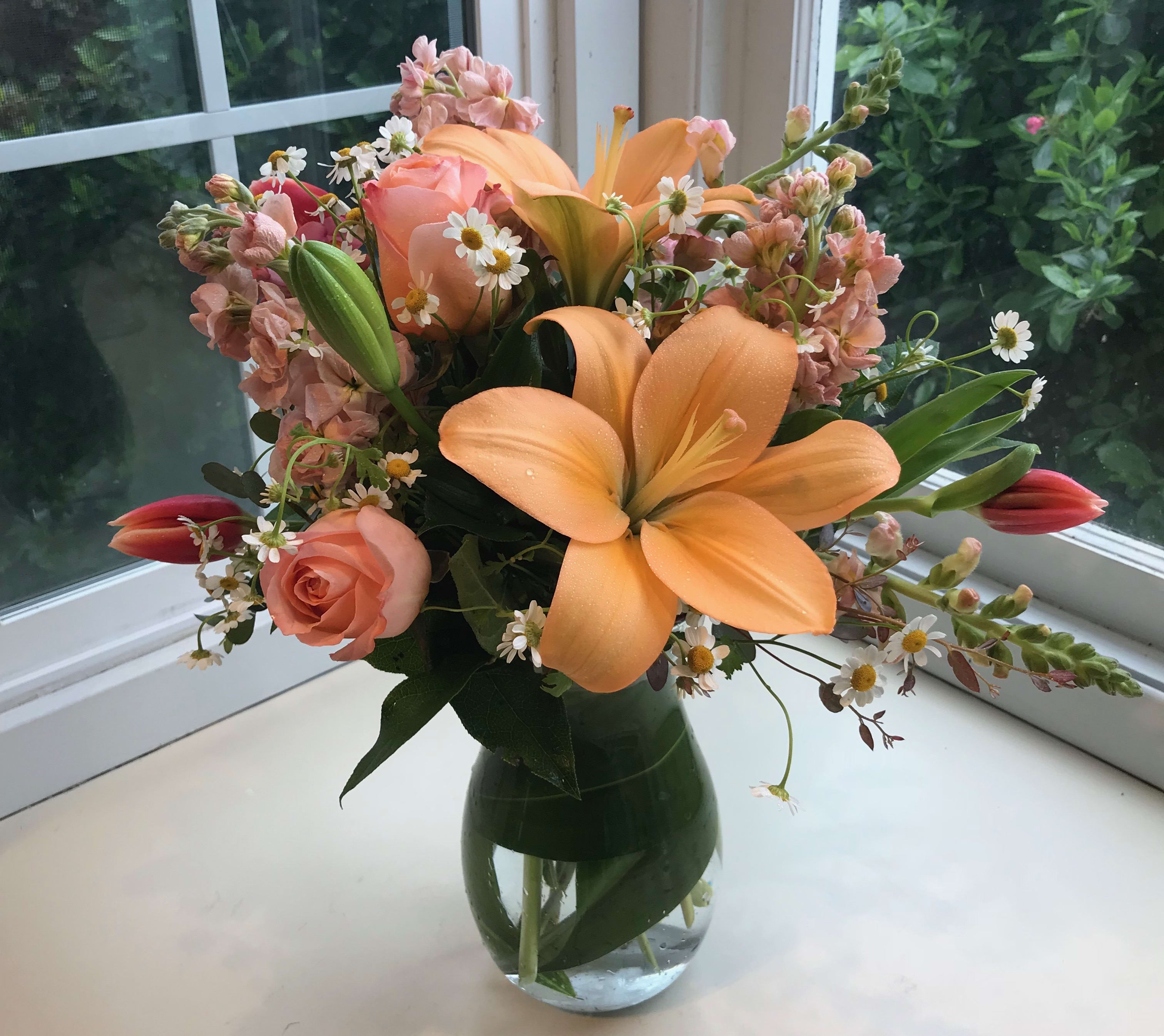 Peachy Keen - Peach lilies, bi-colored roses, stock, and feverfew gather in this display of spring beauty.  A ti leaf is wrapped in the bottom of the vase.  As always, designer may substitute flowers and/or container based on what we have currently in stock.