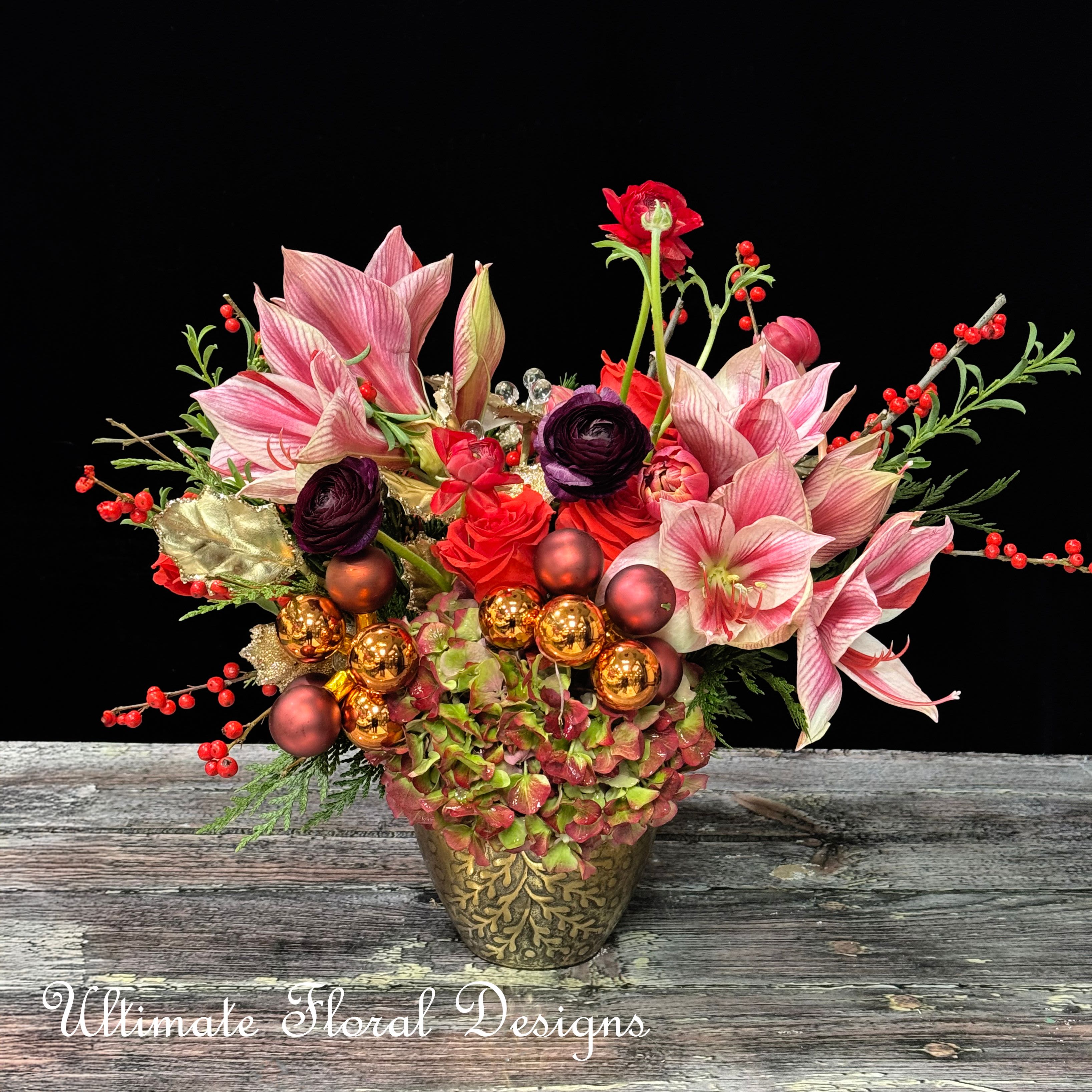 Festive Elegance - This luxurious arrangement exudes holiday sophistication with its stunning mix of pink amaryllis, rich plum ranunculus, vibrant red spray roses, and hydrangeas in earthy tones. Accents of bright red berries and shimmering golden leaves evoke the magic of the season. The addition of metallic ornament spheres adds a contemporary touch, making this arrangement a standout holiday centerpiece. Presented in an ornate golden vase, this design combines traditional elegance with modern flair, perfect for holiday tablescapes or as a lavish gift for someone special.