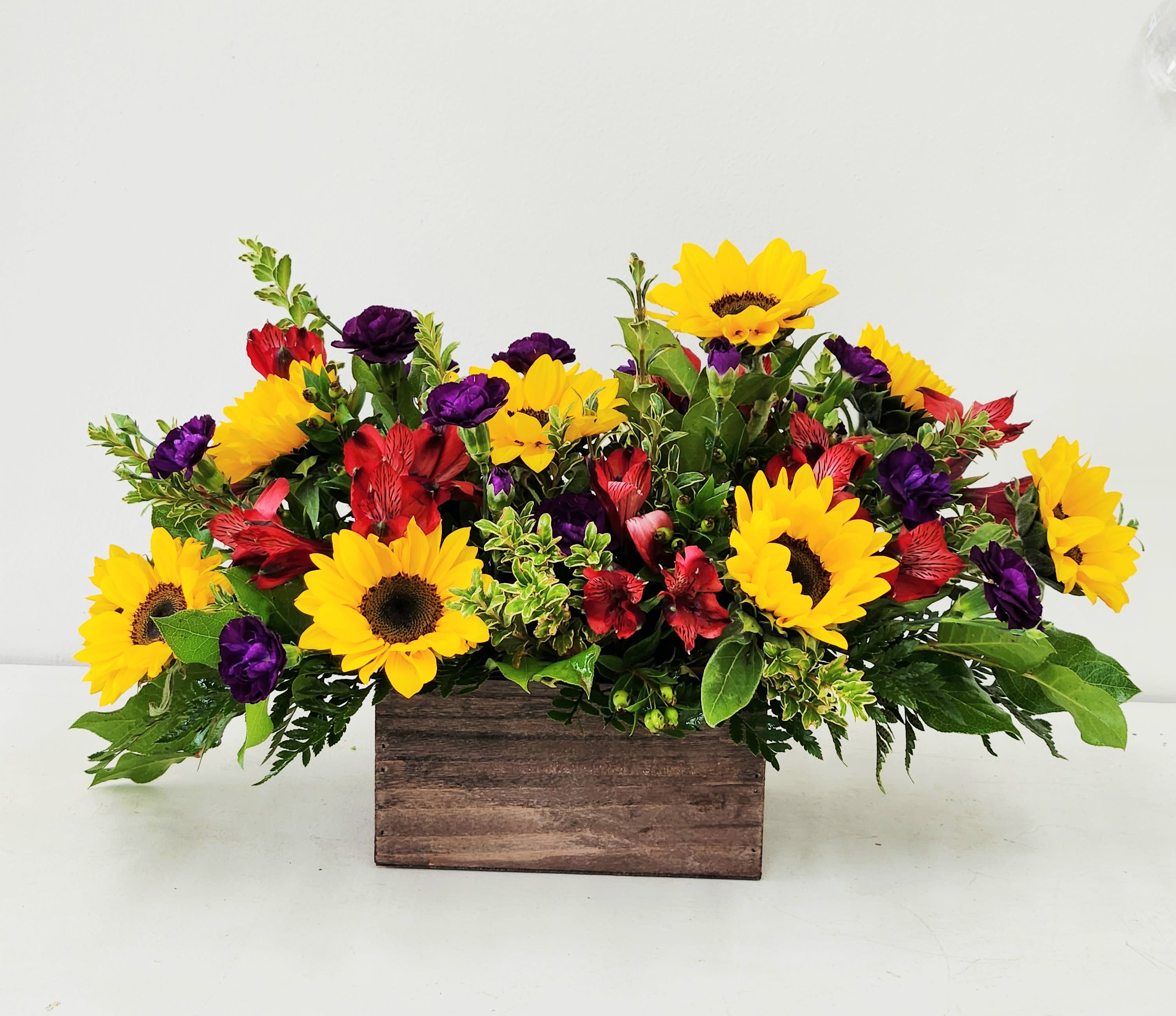 Amber Fields - A cheerful blend of radiant sunflowers, soft spray roses, and vibrant red alstroemerias accented with rich purple carnations — artfully arranged in a rustic wooden box for a warm, country-chic touch.  AS PICTURED: PREMIUM 16&quot;W by 14'H