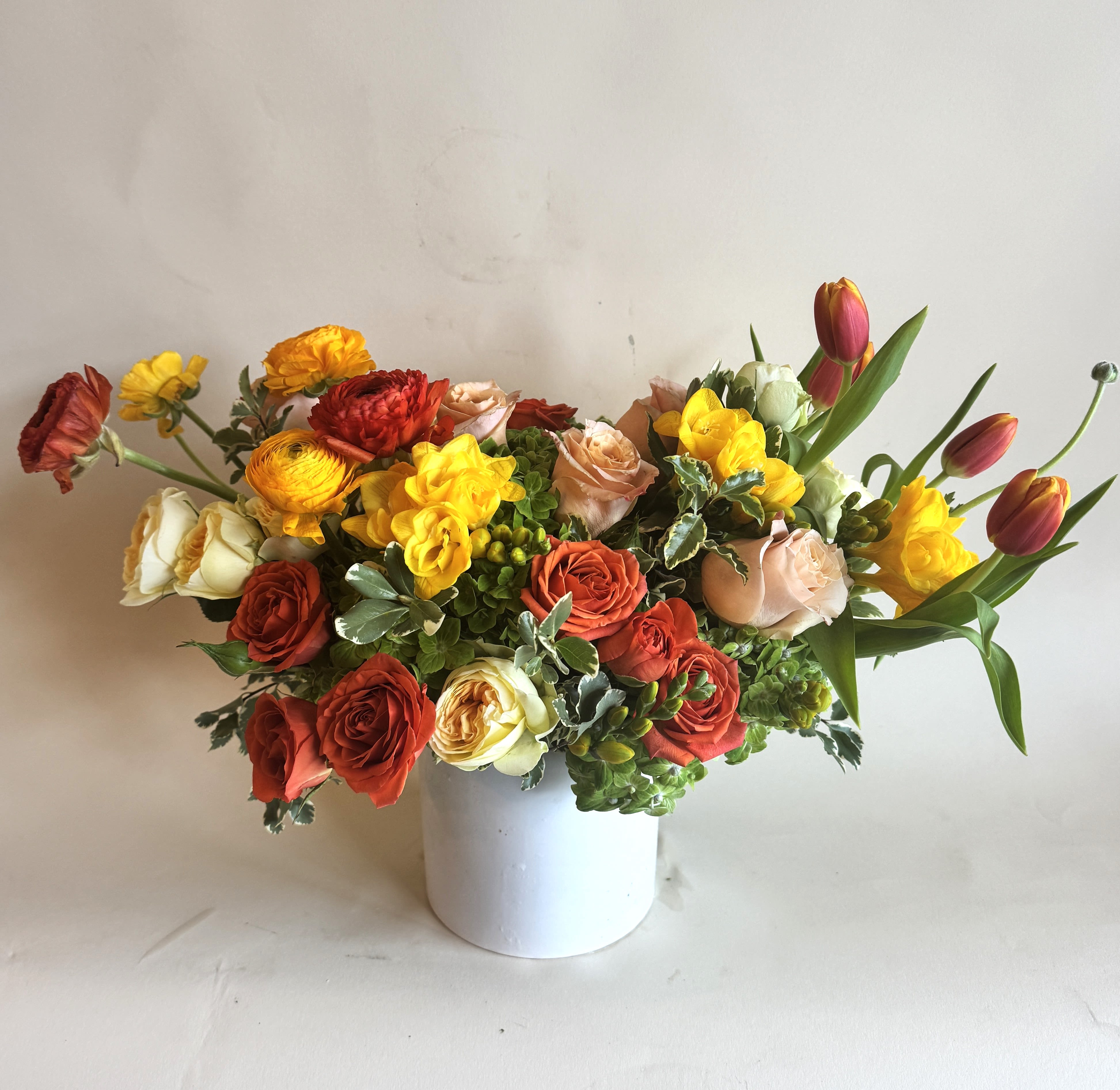 Cherry On Top - A vibrant arrangement with various shades of red, orange and yellow. Arranged in a compact white ceramic vase containing roses, tulips, and ranunculus.  