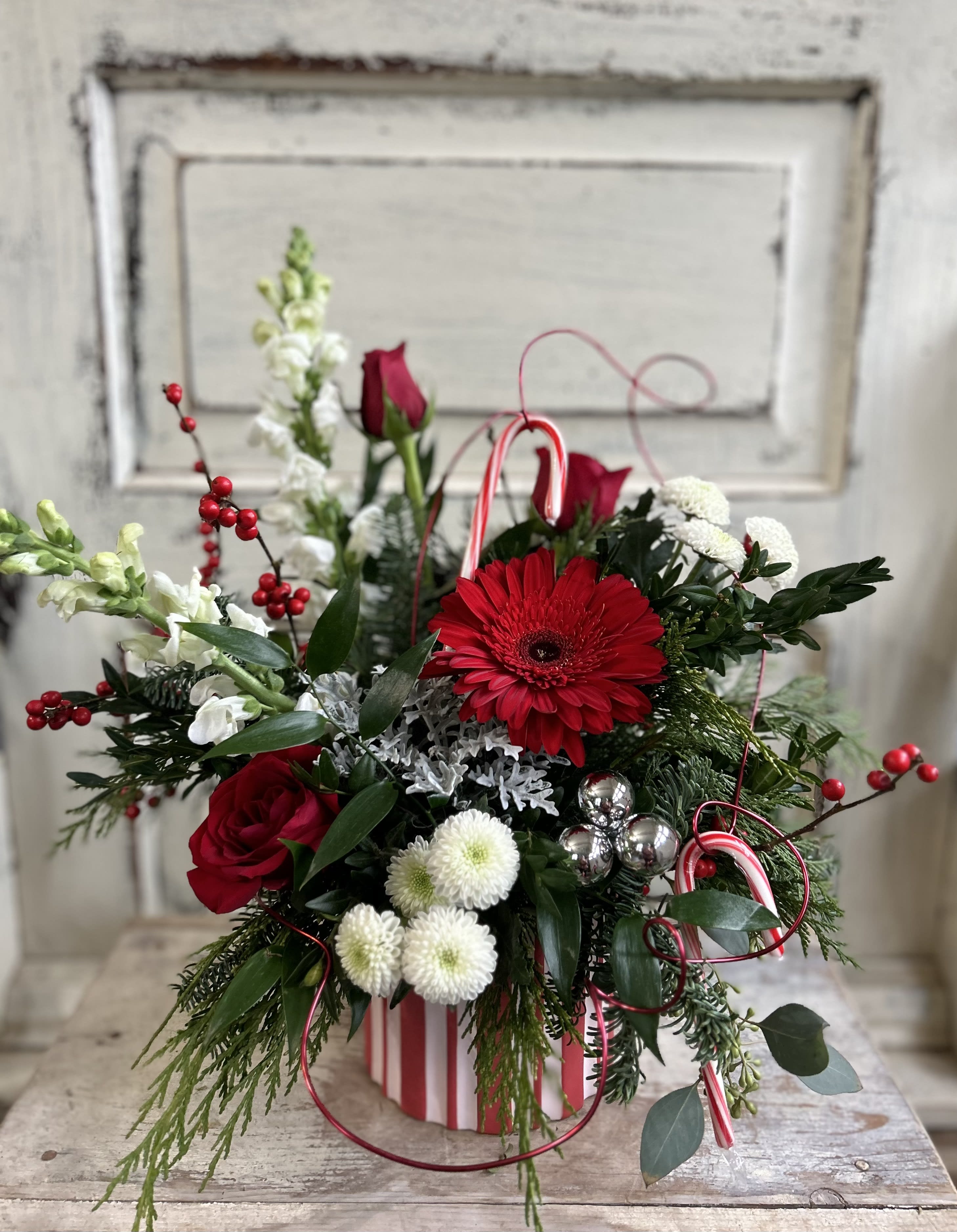Candy Cane Wishes - An adorable red and white candy striped ceramic container is filled with seasonal foliage, red/white flowers, two candy cane treats, and festive trim.