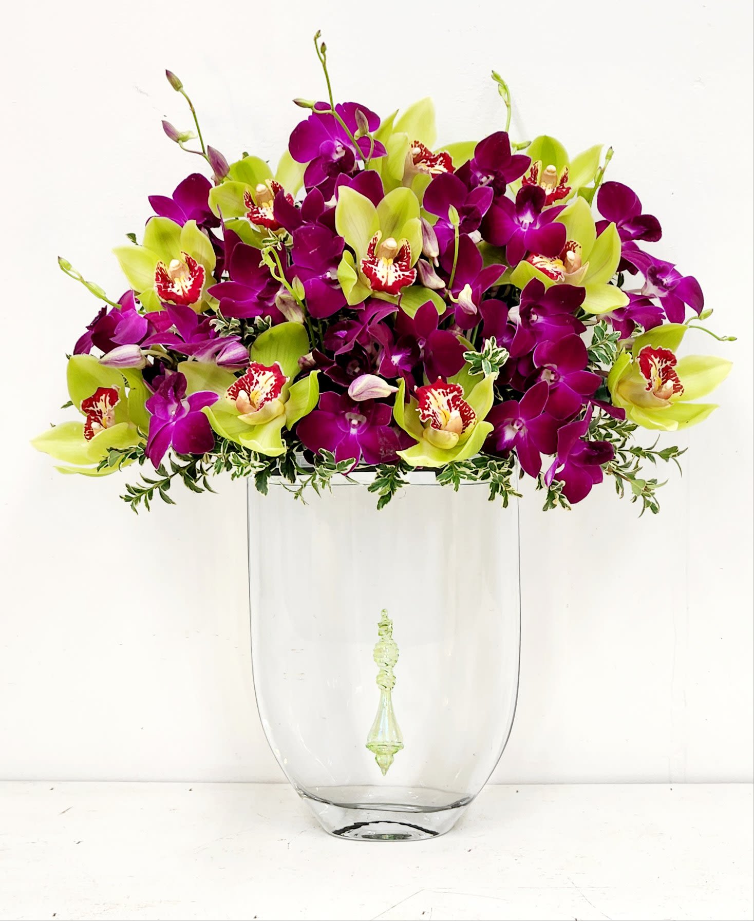 Emerald Ember - A luxurious fall arrangement of green and deep purple orchids, gracefully floating atop a tall clear glass vase. A sparkling pendant suspended below adds a touch of warmth and sophistication, capturing the glow of autumn in a truly one-of-a-kind design. ONE SIZE ONLY. 20&quot;H by 16&quot;W