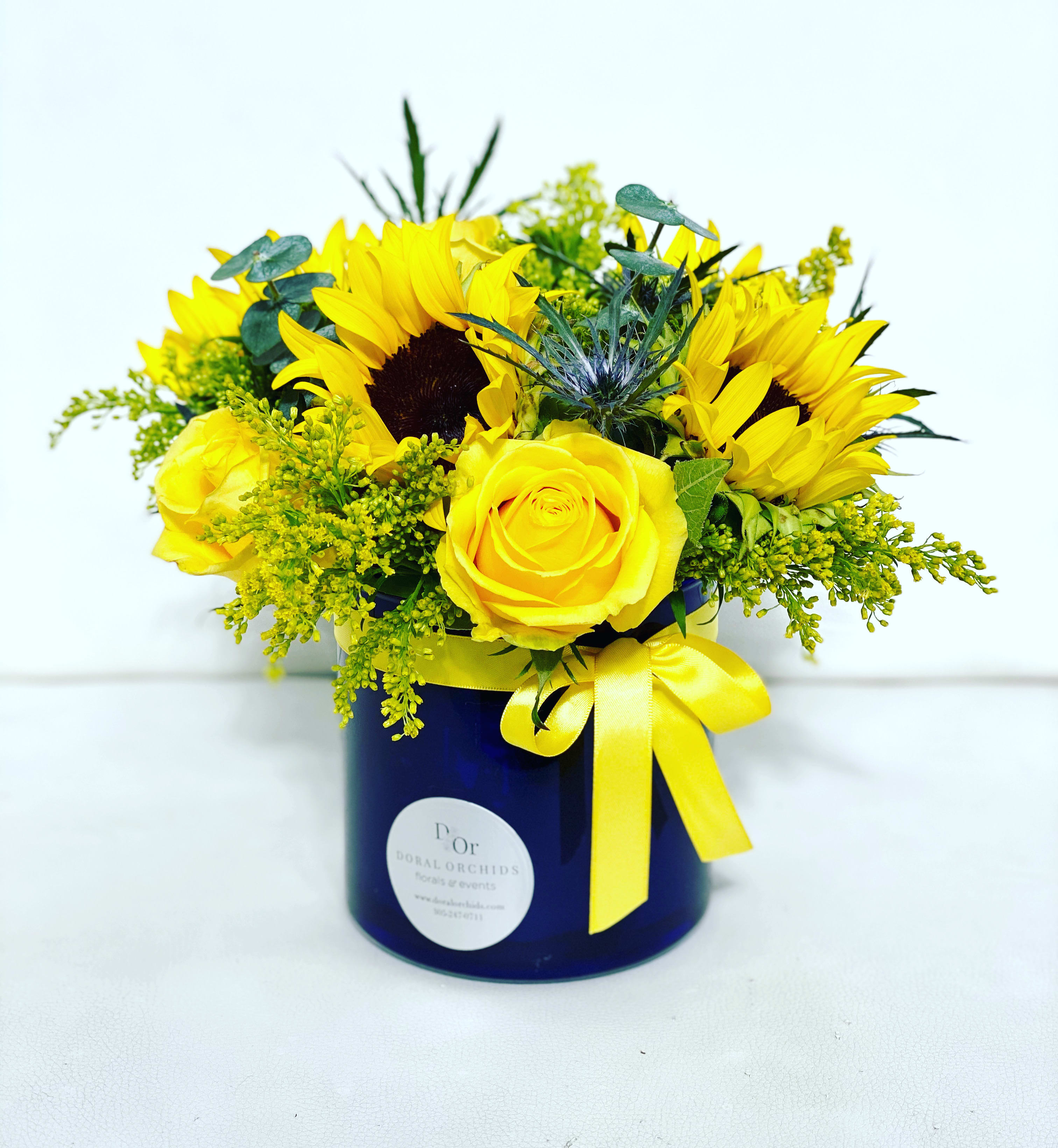Walking on Sunshine ™ - Bright and cheerful, this arrangement beams with golden sunflowers and velvety yellow roses, accented by eucalyptus and playful blue thistle. Designed in a bold navy hatbox with a satin bow, it radiates warmth, joy, and sunny elegance.