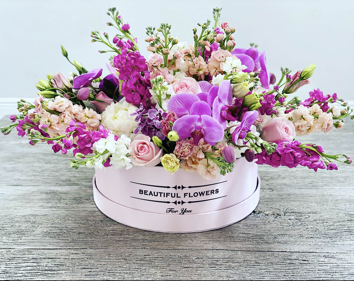 Marvelous ™ - This grand arrangement makes a bold statement, spanning nearly 2 feet wide and about 18 inches tall. Overflowing with exotic purple orchids, lush peonies, and blush roses, it’s accented with sprays of stock and lisianthus for layers of texture and color. Presented in a pastel hatbox, it’s both luxurious and impressively large—perfect as a show-stopping centerpiece.