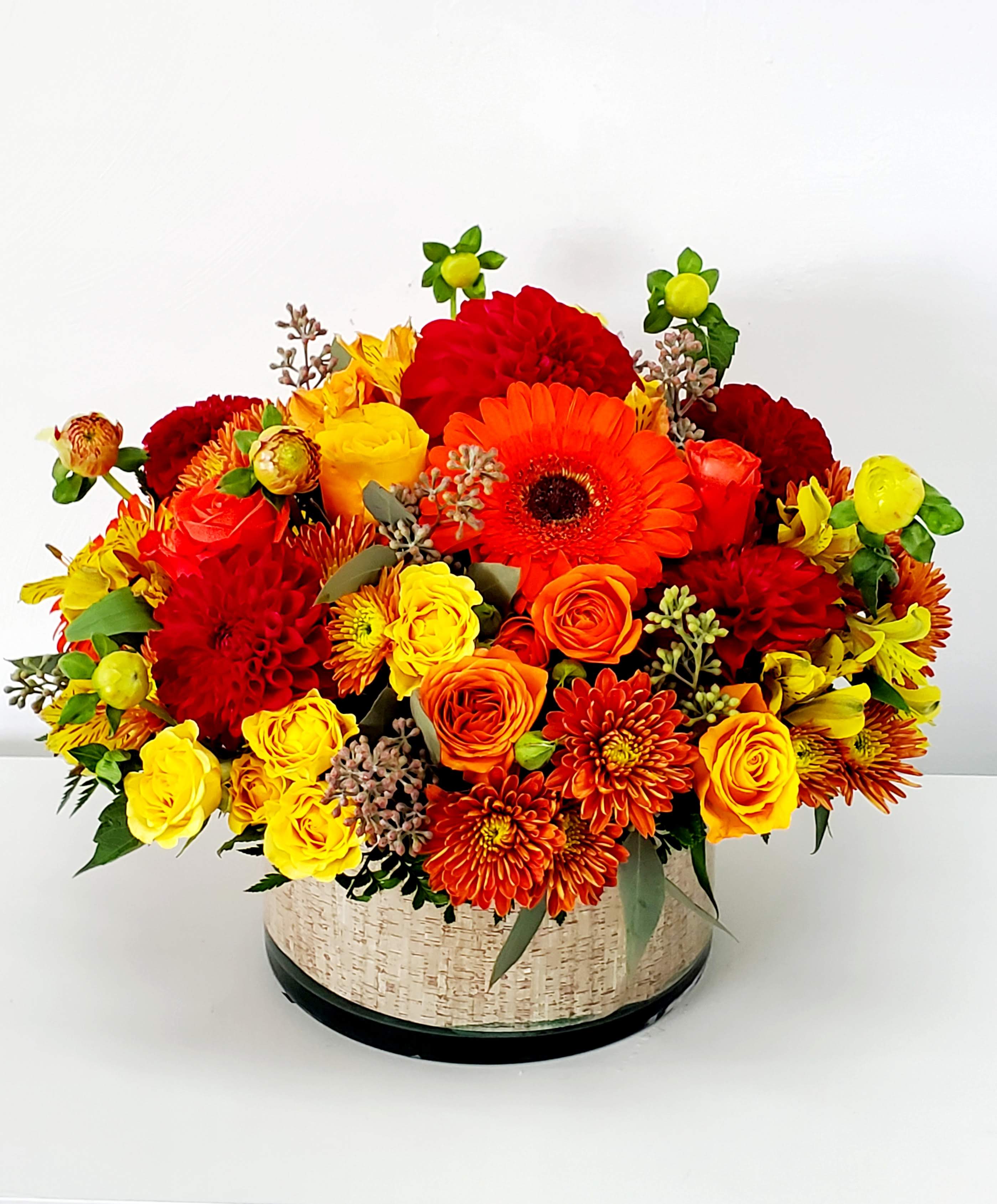 Harvest Glow - This striking centerpiece captures the warmth of autumn. It features an array of red, orange, and yellow blooms arranged in a grounded, wide glass vase. The piece is finished with a unique bark-like lining that adds rich texture, creating a beautiful contrast and making the display a truly glowing focal point for any fall table setting.  AS SHOWN: PREMIUM 16&quot;W x 14&quot;H