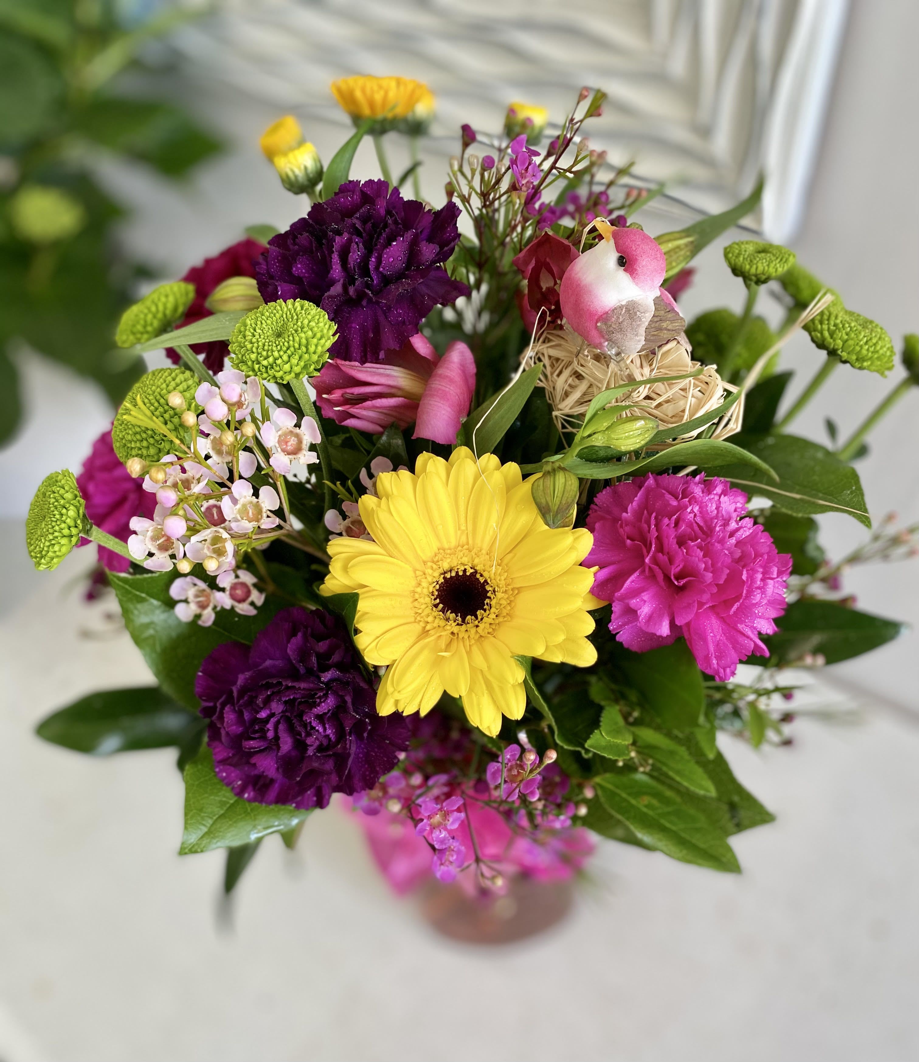 Spring Time  - Add a pop of personality with our Spring Time — a charming bouquet full of bright color and playful detail. Featuring sunny yellow gerberas, rich plum carnations, hot pink blooms, and sprightly green button mums, this petite arrangement is accented with waxflower sprigs and a sweet decorative bird nestled in a cozy straw nest for a touch of whimsy.  Perfect for birthdays, thank-you, or just because, Spring Time brings joy and light-hearted beauty to any moment.