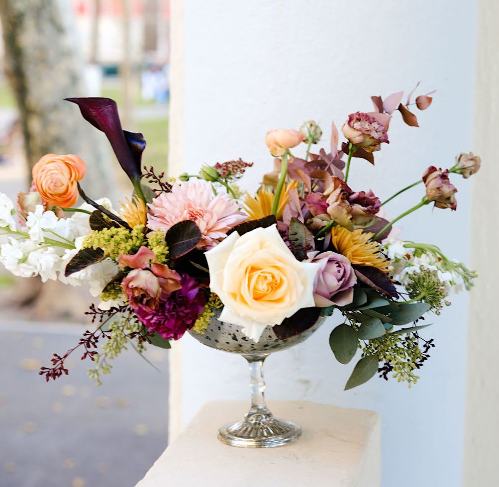 Amethyst Moon - A modern centerpiece designed in a sleek silver compote, featuring dark purple calla lilies, peach and cream roses, and delicate ranunculus. The contrast of moody and soft tones creates a look that’s both sophisticated and effortlessly elegant — a refined statement for the contemporary holiday table.  