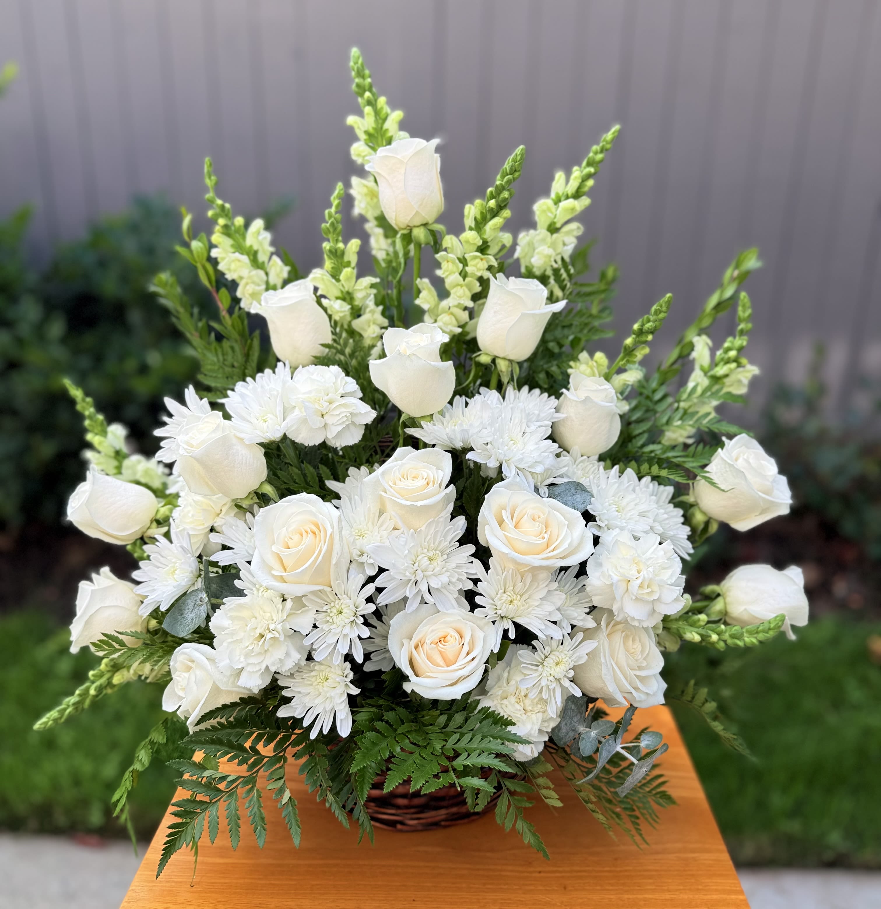 &quot; Serenity &quot; Sympathy Arrangement  - Express your deepest sympathies with this beautiful arrangement that radiates peace; it’s created with harmonious flowers in white and green tones.  It includes: 16 premium roses, snapdragons, cushion pompoms, carnations and seasonal foliage in a rustic basket.  Size is medium 26” H x 23” W One side arrangement. Standard size includes a rustic basket. ——————————————————————- Deluxe size includes : roses, snapdragons, Pompoms, campanula, white little flowers, seasonal greenery in a white vase .