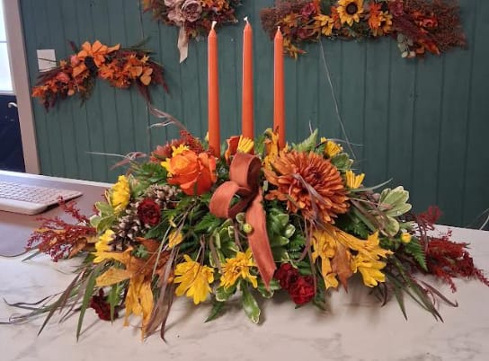 Triple Candle Centerpiece - Set the scene for celebration with this triple-candle centerpiece, filled with the warmth of the season. Featuring a blend of autumn flowers, pinecones, and colorful fall leaves, it brings a welcoming glow and a touch of natural beauty to your holiday table.
