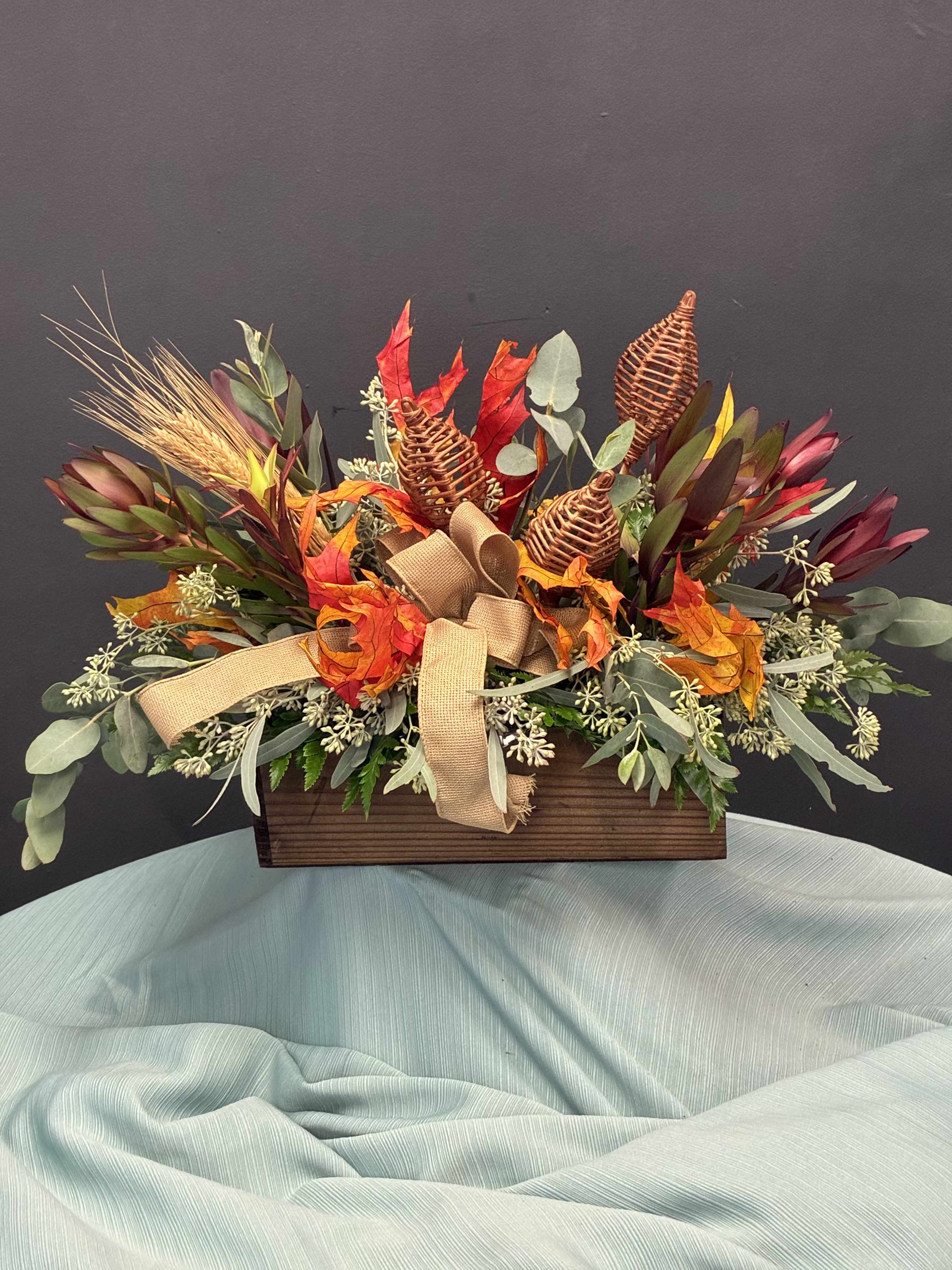 Harvest Keepsake  - A festive blend of autumn’s finest textures and tones. Vibrant red, orange, and yellow blooms mingle with dried wheat, Saffari accent , and lush greenery, all nestled in a rustic wooden box. A burlap bow adds a homespun charm, making this arrangement a warm invitation to gather, give thanks, and celebrate the season’s bounty. Perfect for home decor, Offices, Lobbies and much more!