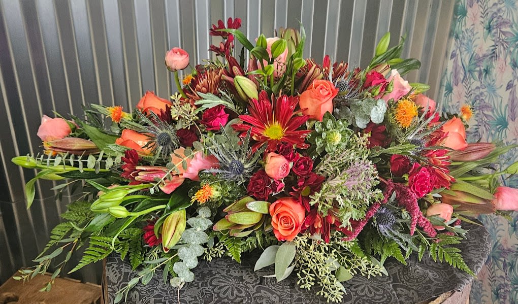Crimson Harvest Elegance centerpiece - Make a statement with our Crimson Harvest Elegance centerpiece — a luxurious fall floral design overflowing with roses, lilies, chrysanthemums, thistle, and lush greenery. Deep crimson tones mingle with soft peach and orange hues for a breathtaking autumn presentation full of texture, movement, and natural beauty. Designed for dining tables, events, memorials, or seasonal décor, this arrangement captures the richness of fall in every petal. Handcrafted by skilled local florists using only the freshest premium blooms, this piece adds warmth and elegance to any setting. Available for same-day delivery if ordered by 12 noon, and custom size upgrades also available.