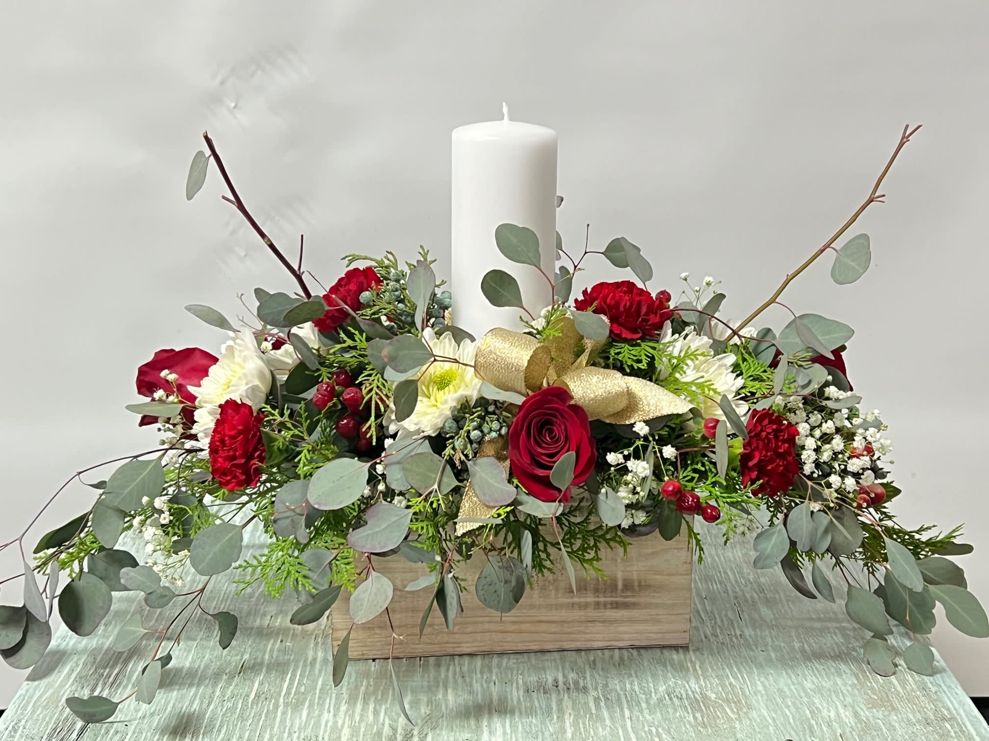 Christmas #10 - An arrangement of fresh cut flowers designed in a wooden box container. Embellished with gold ribbon and a candle in the center. Traditional centerpiece style.