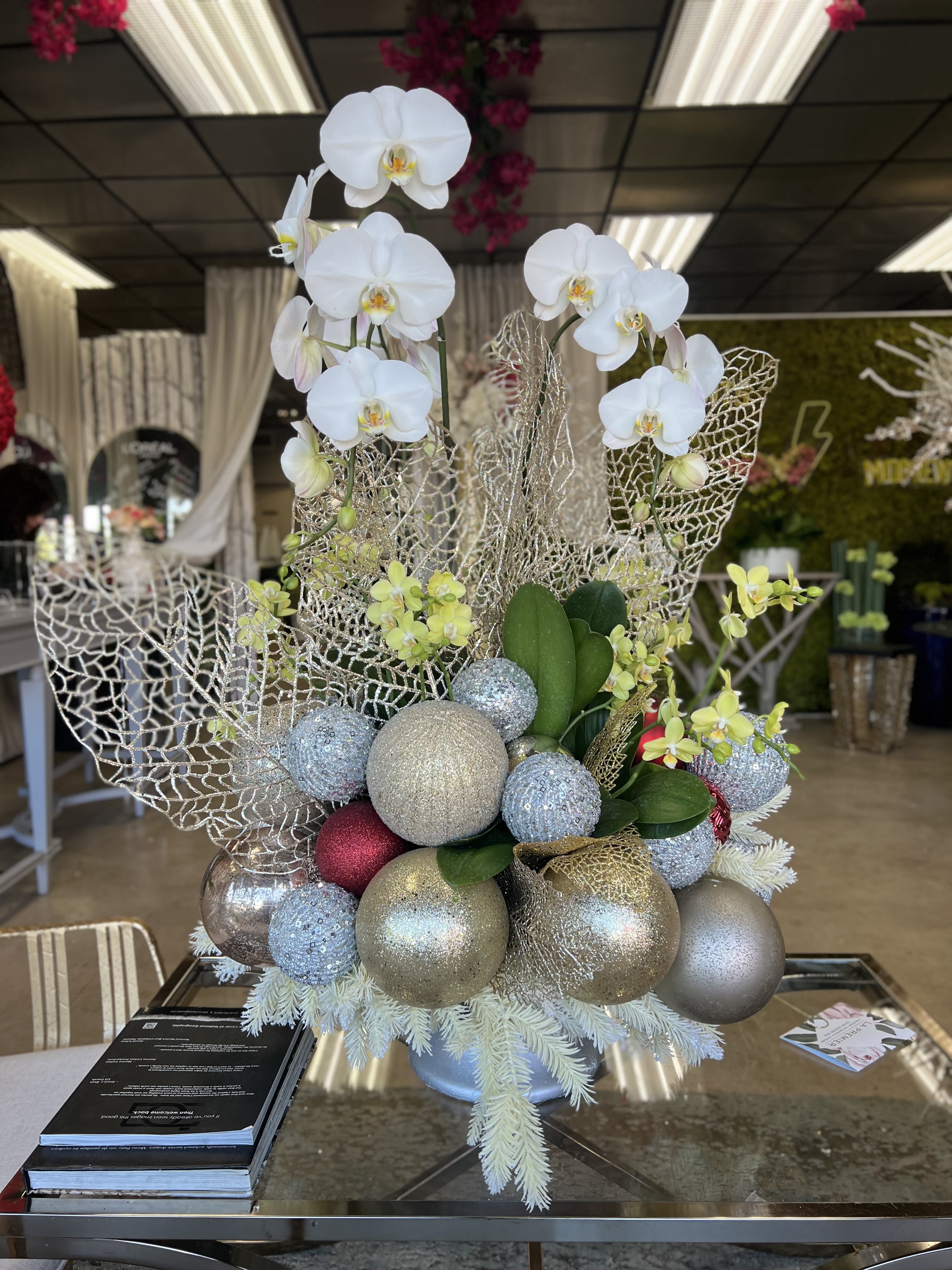 Jingle Bells - Introducing the perfect holiday centerpiece – a beautiful Christmas Orchid Arrangement that radiates elegance and festive charm. This stunning piece features a delicate blend of large white orchids and vibrant yellow baby orchids, creating a striking contrast that embodies the spirit of the season. Accentuated with festive golden ornaments, this round arrangement captures the warmth and beauty of Christmas, making it the ideal decoration for your home, office, or holiday gatherings.  The pure white orchids symbolize peace and purity, while the cheerful yellow baby orchids add a touch of brightness to your celebration. Adorned with sparkling ornaments, this centerpiece is a true work of art – bringing sophistication and joy into any room. Whether you're gifting someone special or enhancing your holiday décor, this orchid arrangement is the perfect way to bring the magic of Christmas to life.  Make your holiday season unforgettable with this exquisite, one-of-a-kind floral masterpiece.