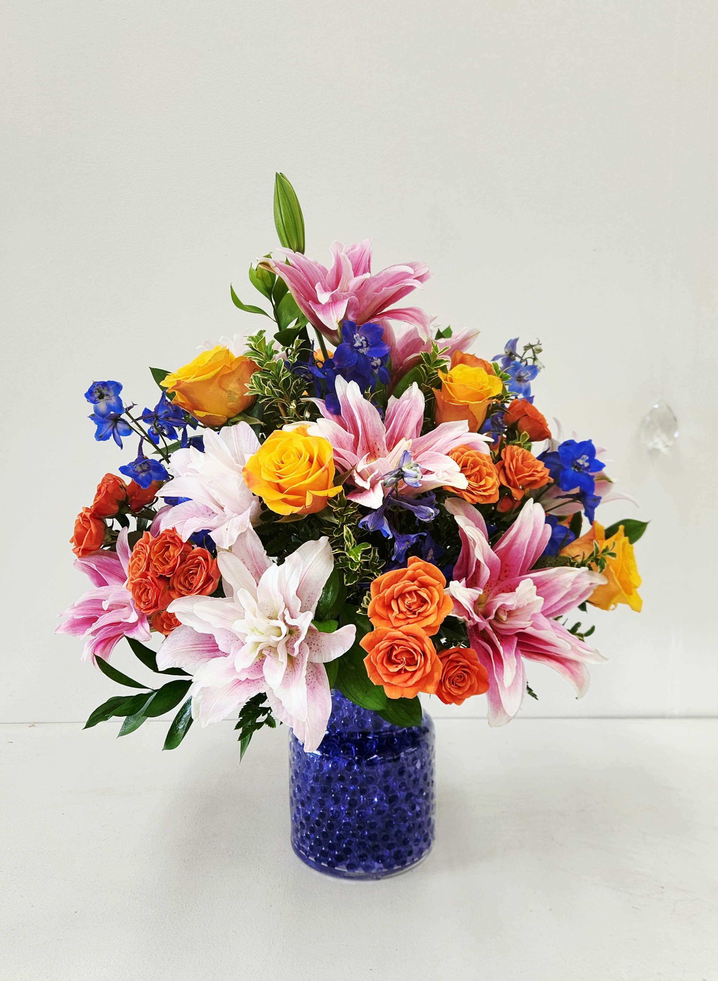 Gorgeous! Like You - Turn heads with this show-stopping bouquet bursting with vibrant orange, pink, and blue lilies alongside sunny yellow blooms. handcrafted in a glass vase filled with Blue Water Gels. Uniquely different! It's the kind of arrangement that makes people stop and say &quot;wow&quot;—luxurious, bold, and unapologetically gorgeous. Whether you're celebrating a milestone, saying thank you, or just want to brighten someone's world, this colorful stunner delivers major joy.&nbsp; ONE SIZE ONLY 18&quot;W by 22&quot;H