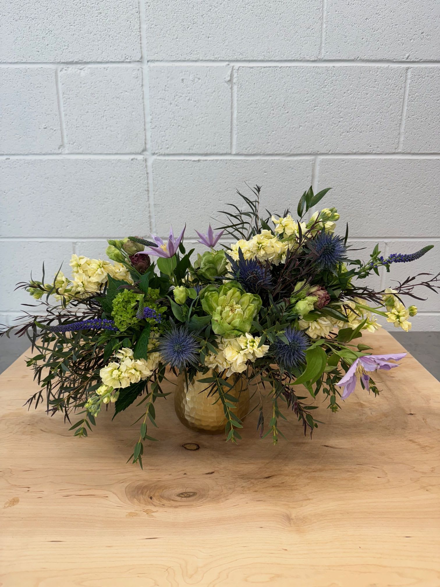 Autumn's Whimsy - This airy arrangement is full with Agonis, Gunnie Eucalyptus, Globe Thistle, Pale Yellow Stock, Veggie Roses, Clematis, and Purple Veronica.  The container is a water tight hammered gold tin.  Finished, the arrangement is roughly 18&quot; Wide and 12&quot; Tall.  Long lasting, with a light scent.