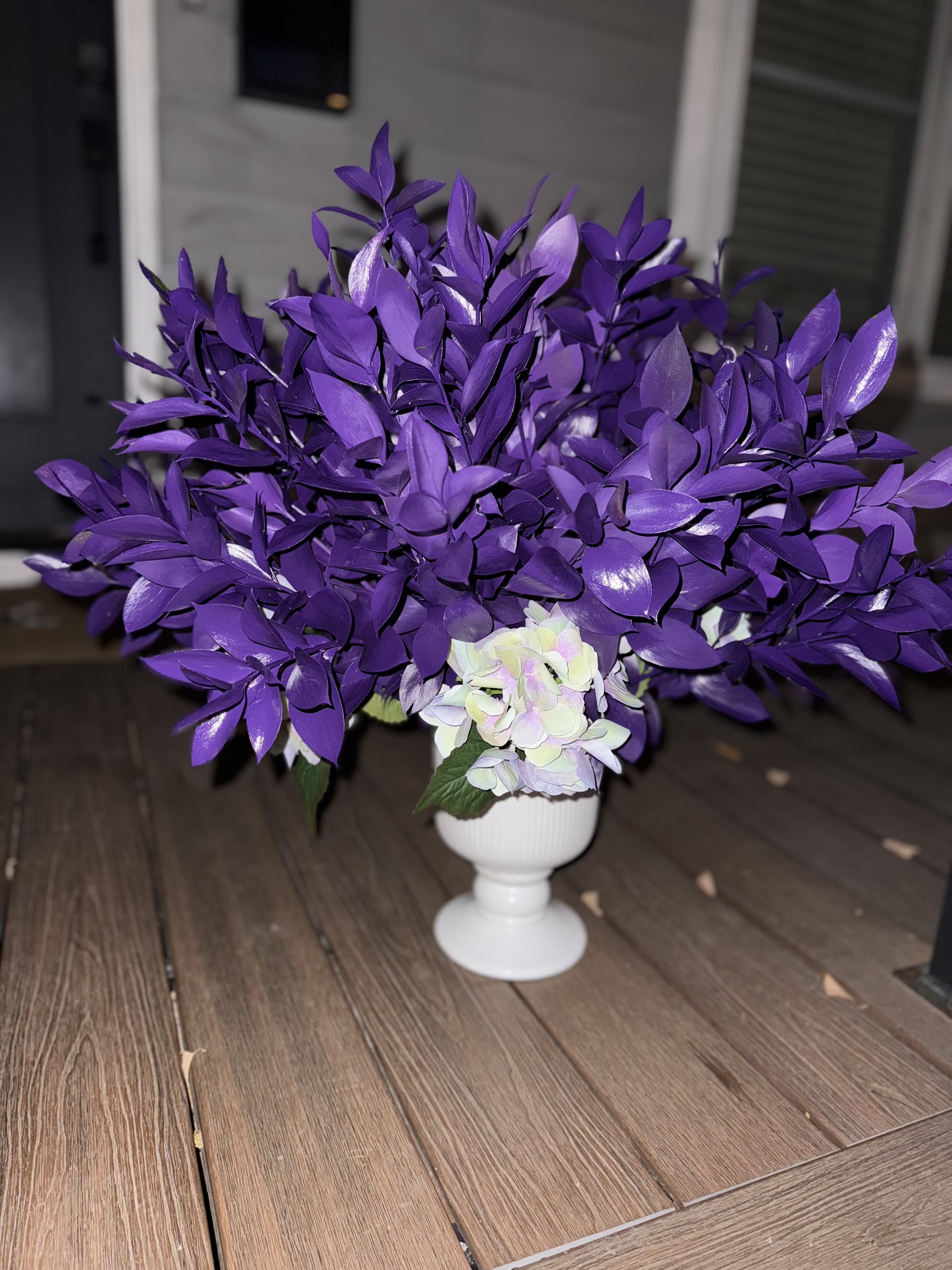 Royal Radiance - A statement piece that embodies elegance and power — the Royal Radiance arrangement features deep, velvety purple foliage paired with soft hydrangeas for a touch of grace. Designed in a sleek white pedestal vase, this bouquet is perfect for making bold entrances at luxury events or adding a touch of opulence to intimate spaces. Symbolizing strength, creativity, and divine favor, this arrangement is a reminder that standing out is always in season.