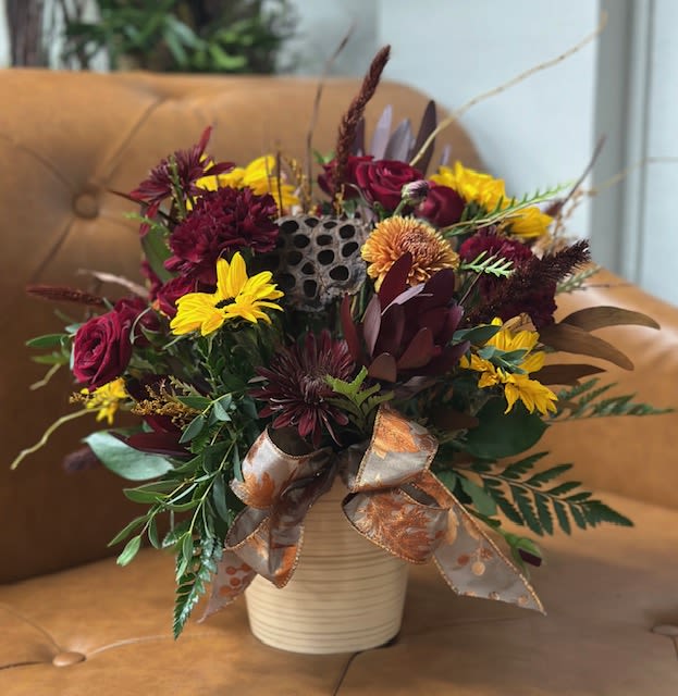 Burgundy Harvest Charm - This rich and radiant arrangement captures the heart of fall with deep, velvety tones and warm golden accents. Burgundy spray roses and mums pair beautifully with vibrant mini sunflowers, Textured elements such as the dried lotus pod, preserved foliage, and rustic cattails, bring an earthy, woodland feel. Nestled in a neutral ribbed ceramic container and finished with a shimmering autumn ribbon, this centerpiece radiates cozy seasonal elegance.