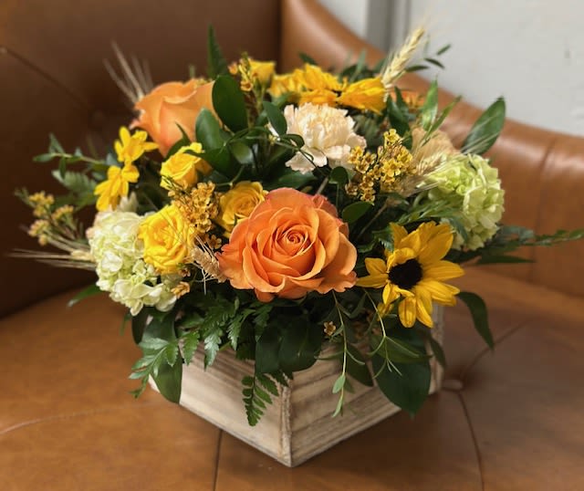 Country Sunflower Crate - Bright, cheerful, and full of harvest warmth, this arrangement brings the glow of an autumn sunrise indoors. Lush orange roses sit and the heart of the design, complemented by sunny yellow spray roses, mini sunflowers, and mini green hydrangeas. Wheat stems and seasonal greenery lend a rustic, farmhouse touch. Designed in a distressed wooden box.