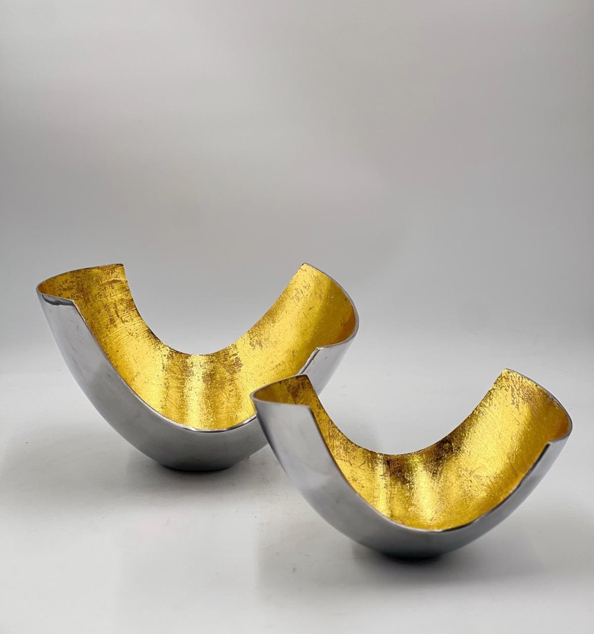 Golden Arc Bowls - These modern and sophisticated decorative bowls feature a sleek silver exterior with a stunning gold-leaf interior, creating a striking contrast that exudes elegance and luxury. The curved, asymmetrical shape adds a contemporary touch, making them versatile accent pieces for any home. Perfect for holding small decorative items, serving as a centerpiece, or standing alone as a statement piece, these bowls are ideal for elevating any space with their chic design. The price includes both items, making it a perfect set for gifting or personal use.