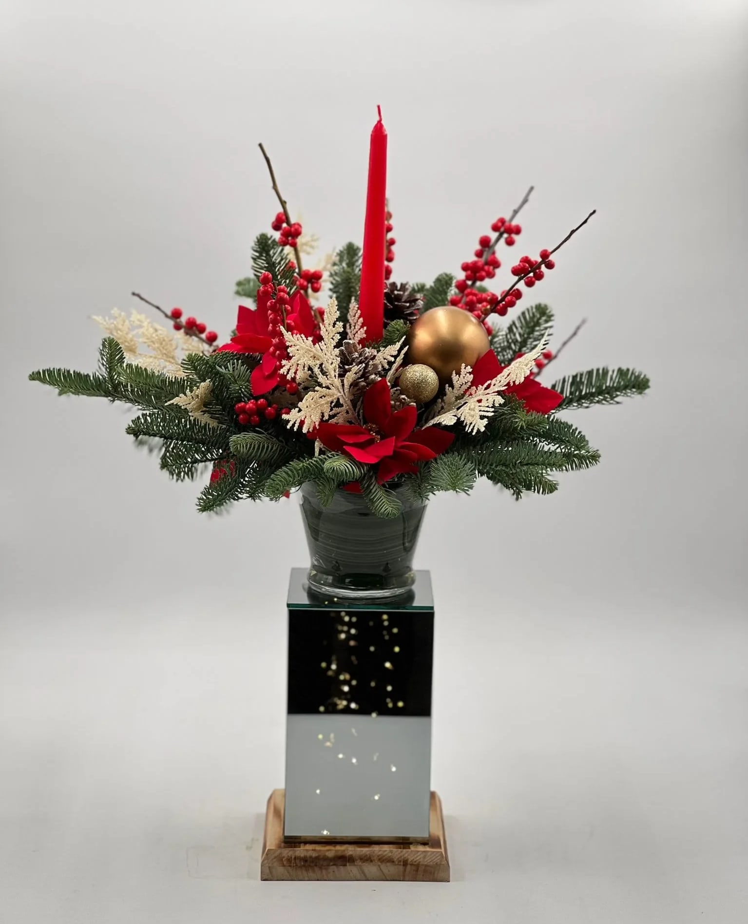Winter Wonder - This is a festive holiday centerpiece featuring a vibrant arrangement of seasonal foliage and decorations. The centerpiece includes lush evergreen branches, punctuated by rich red flowers and clusters of holly berries. Accented with gold and red baubles and dusted with snow-like speckles, the arrangement creates a warm and inviting holiday atmosphere. The focal point is a tall, elegant red candle that promises to cast a soft, ambient glow. The arrangement is presented in a stylish, reflective vase mounted on a wooden base, adding a contemporary touch to the traditional festive decor. The light within the container adds a subtle illumination, enhancing the arrangement's allure and bringing a soft, festive glow to the surroundings. 