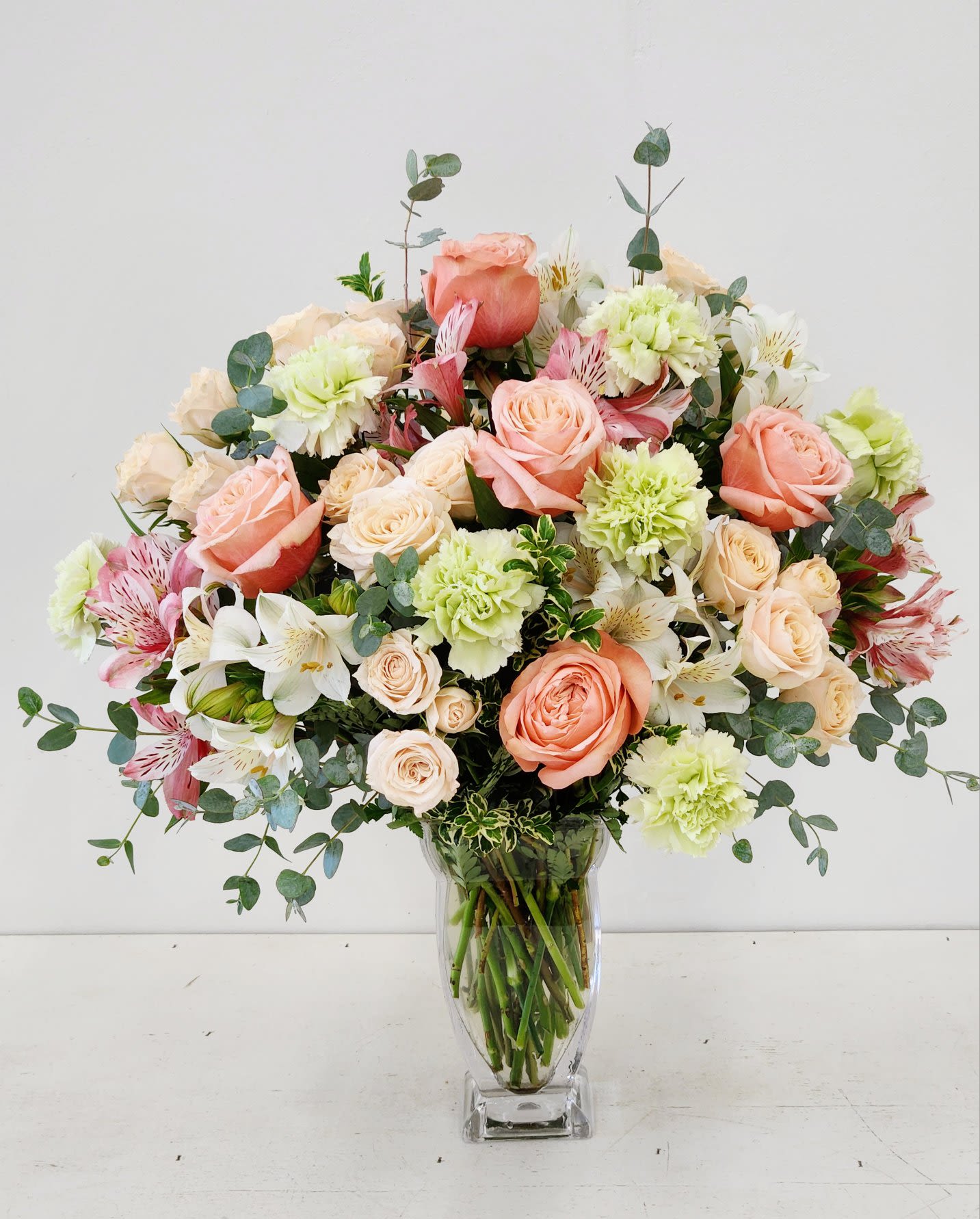 Peaches and Green - Soft peach roses and green carnations blend seamlessly with delicate peach spray roses and pristine white alstromerias in this refined arrangement. Graceful eucalyptus greenery adds an elegant finishing touch, creating a sophisticated composition that balances warmth with natural beauty. Perfect for celebrating life's special moments with understated luxury.