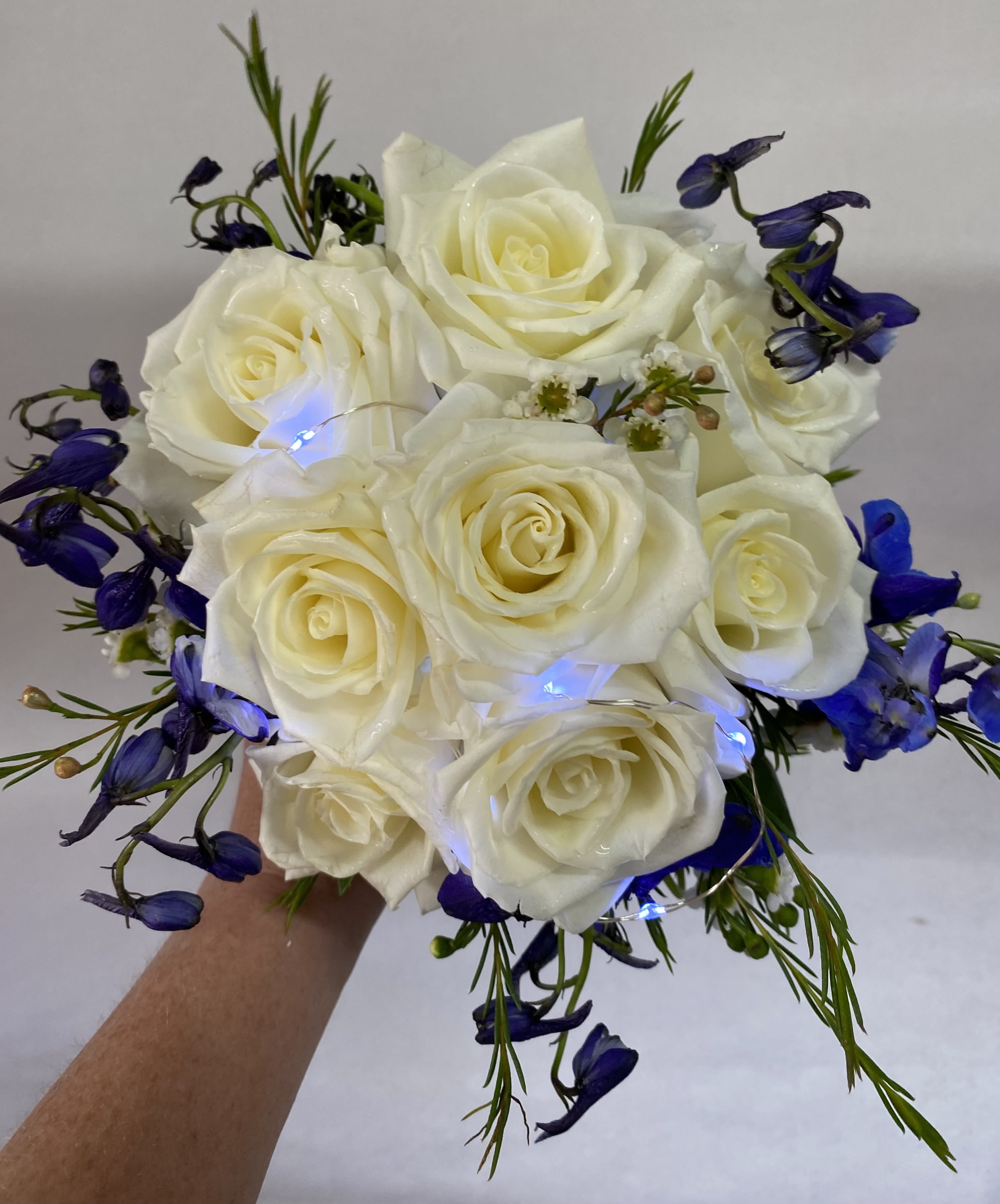 Sophia's Fairy Lights Hand Held Bouquet - White roses with blue delphinium with fairy lights for a magical glow. Fairy lights come in white and soft gold. Please give us a call if you would like different colored flowers.