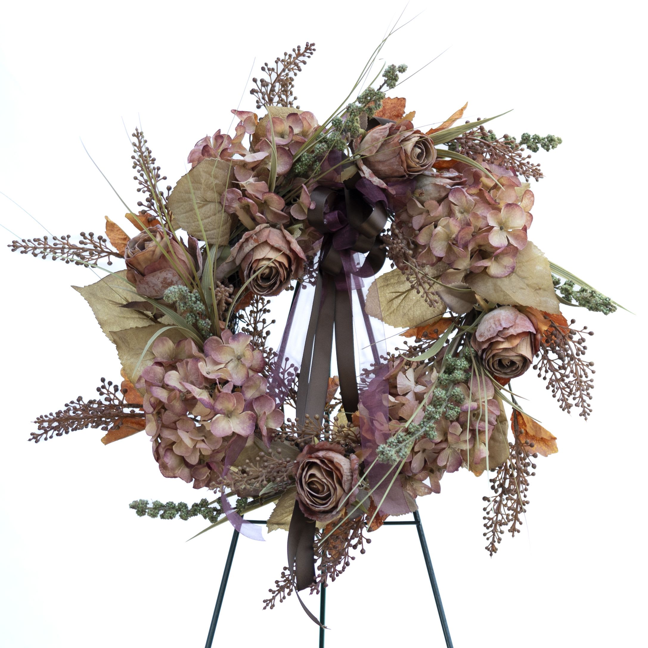Autumn Serenity Wreath - A floral wreath, designed for decorative or memorial purposes. The wreath features a mix of dried or artificial flowers, including hydrangeas and roses, along with various foliage and accent pieces like grasses and seed pods. The colors are soft, dominated by muted shades of pink, beige, and brown, giving the arrangement a rustic, autumnal feel. A dark ribbon is tied at the center, adding a touch of elegance to the overall design.