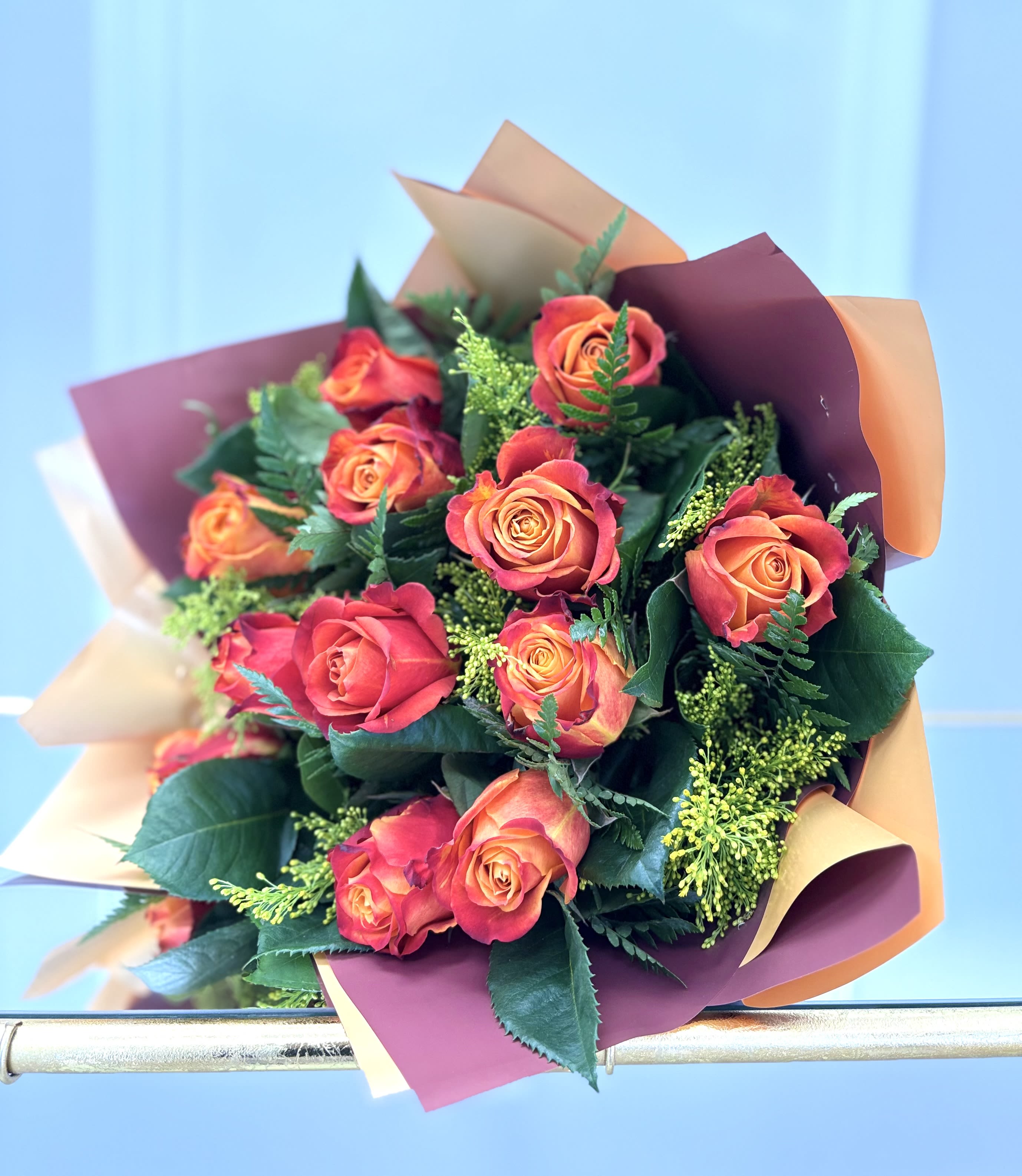 Firelight Kiss By VLM Flower - Radiating warmth and passion, Firelight Kiss is a striking bouquet of sunset-hued roses wrapped in deep amber and burgundy tones. Each rose blooms with fiery gradients of orange, coral, and red, paired with lush greenery and delicate golden filler to enhance its rich glow. Bold yet elegant, this bouquet is perfect for celebrating love, anniversaries, or simply igniting someone’s day with a burst of vibrant beauty.