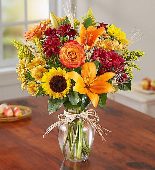 Autumn Fields - All-around arrangement with autumn-colored roses, sunflowers, orange Asiatic lilies, red and autumn-colored daisy poms, accented with wheat, solidago and assorted greenery. Artistically designed in a clear glass vase accented with raffia. Arrangements are hand-designed so colors and varieties may be substituted with similar items based on local availability. 