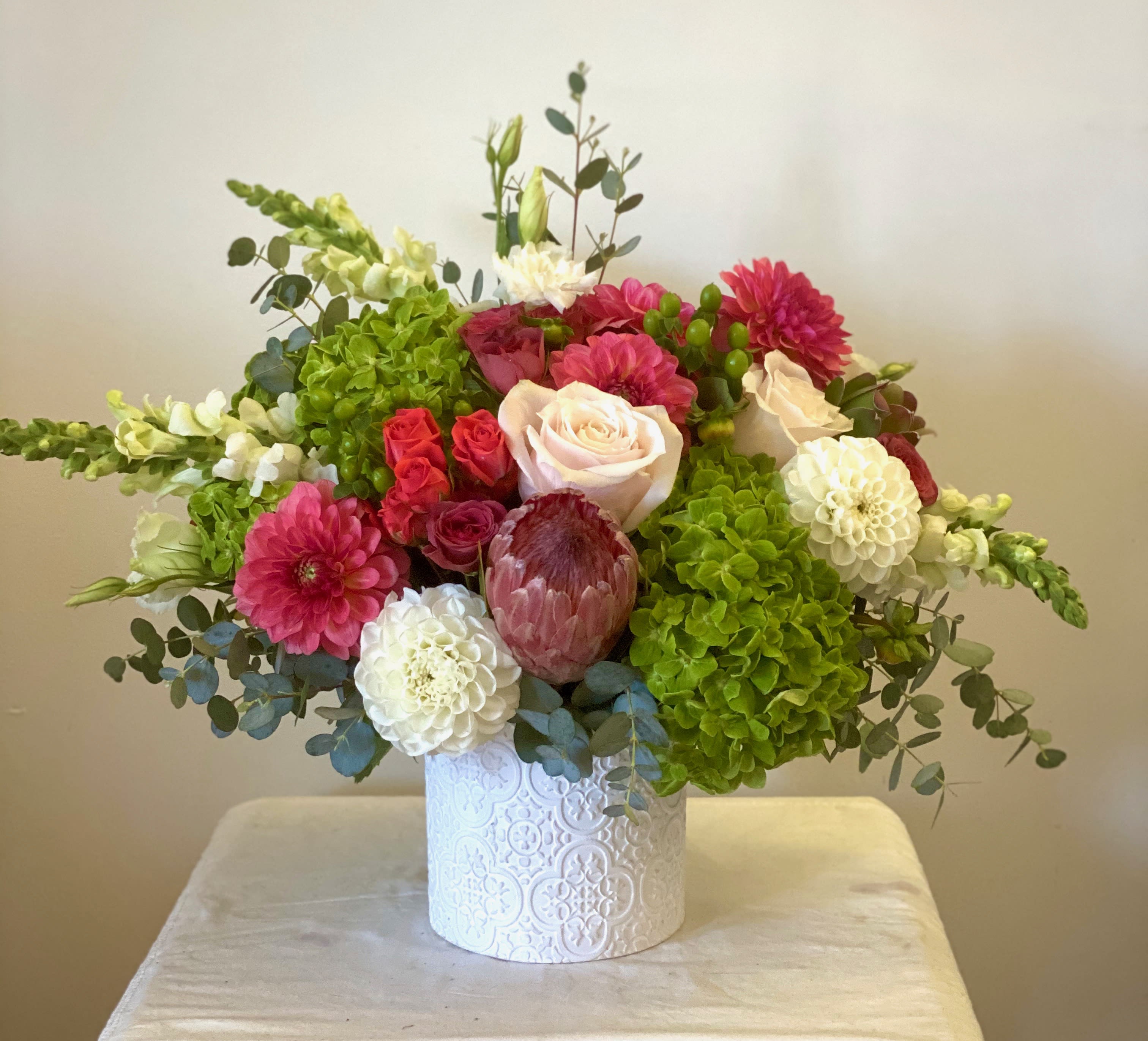 Ruby's Little Gems - Seasonally, whichever Protea is in season we will use the first photograph is pink mink Protea, and that will include the following-Coral or red roses, pink mink protea, dahlias, lisianthus, green hydrangea, white snapdragons and greens in a unique ceramic vase. Pincushion Protea typically comes in orange or yellow, and the colors will coordinate and lastly, when no protein is available, we use Banksia and coordinating roses and seasonal blooms.