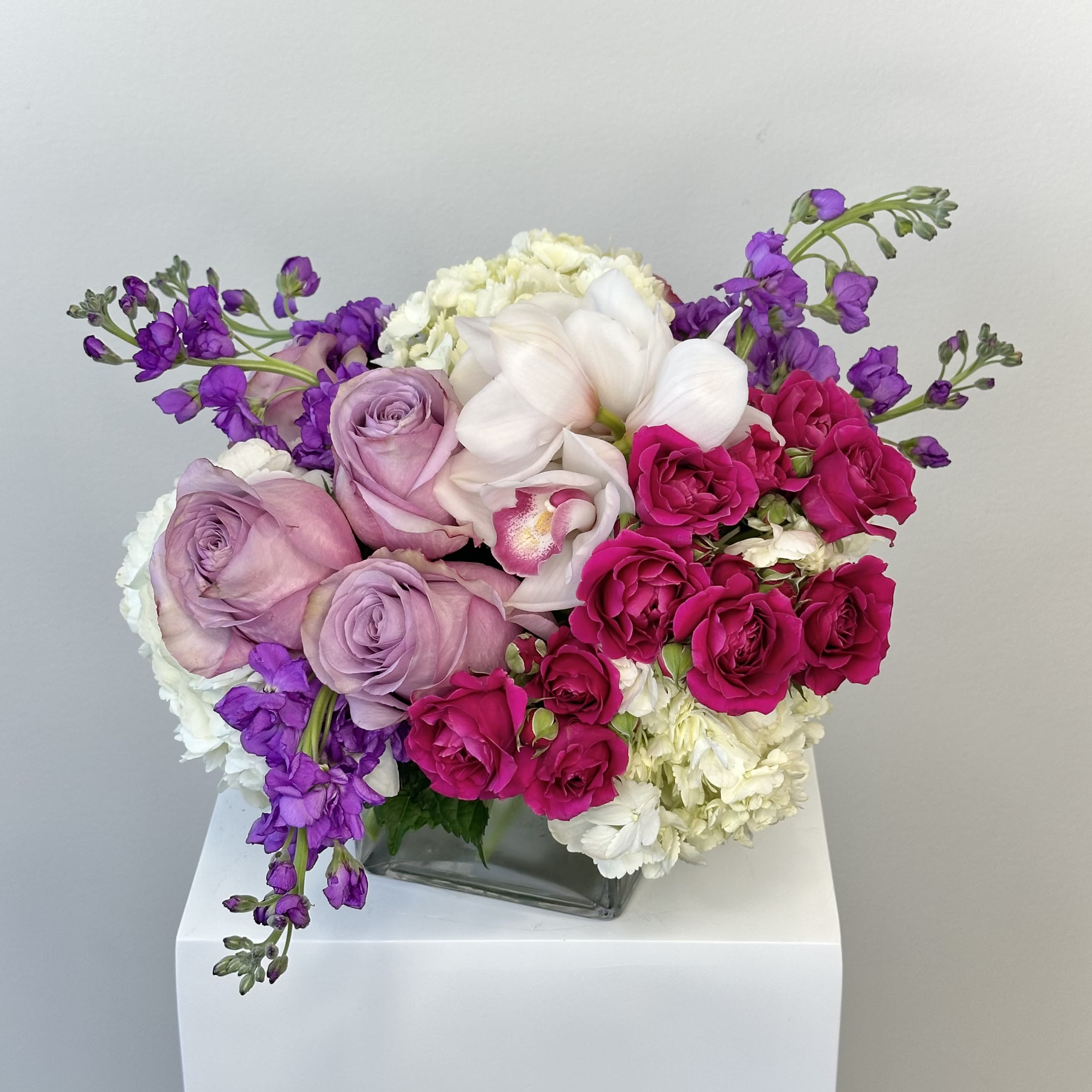 Velvet Wave - Drift into elegance with the Velvet Wave arrangement, a stunning cascade of white hydrangea, purple roses, vibrant hot pink spray roses, fragrant purple stock, and exotic white cymbidium orchids. This opulent bouquet blends rich textures and jewel tones for unforgettable impact—perfect for anniversaries, birthdays, sympathy, or luxury décor.  Why Choose Velvet Wave? Expertly designed with plush white hydrangea for abundance, velvety purple roses symbolizing enchantment, playful hot pink spray roses for joy, aromatic lavender/purple stock evoking nostalgia, and long-lasting white cymbidium orchids for refined beauty. Arrives in a premium vase or hand-tied with satin ribbon. Ideal for modern wedding flowers, romantic gifts, or corporate events.  Bloom Symbolism: White Hydrangea: Gratitude, purity Purple Roses: Love at first sight, majesty Hot Pink Spray Roses: Appreciation, fun Purple Stock: Lasting beauty, contentment White Cymbidium Orchids: Luxury, strength  Search velvet wave flowers, purple roses white hydrangea bouquet, hot pink spray roses stock orchids, lavender purple floral delivery, or luxury cymbidium arrangement? Velvet Wave ranks high for designer sympathy bouquets, same-day premium flowers, anniversary orchid gifts, and jewel-tone arrangements.
