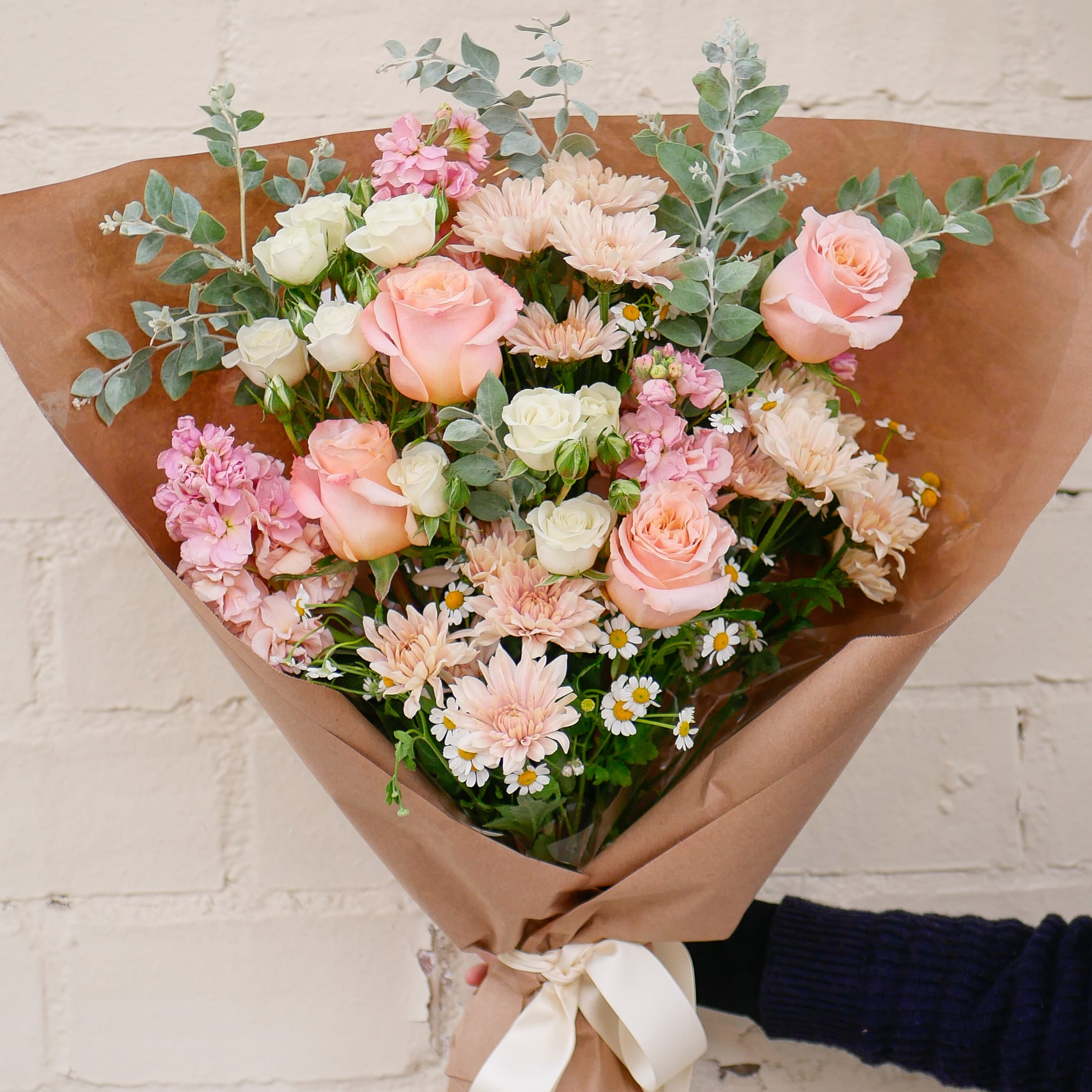 Pretty In Peach Bouquet - This pastel bouquet displays a gorgeous mix of peach and white blooms, including roses, mums, stock and chamomile.  NOTE: This bouquet DOES NOT include water tubes or a vase. It is wrapped with cellophane and brown paper and needs to be placed in water immediately after receiving. The design is &quot;presentation&quot; style as a flat lay to make a floral statement for the recipient, it will need re-arranging when placed in a vase.