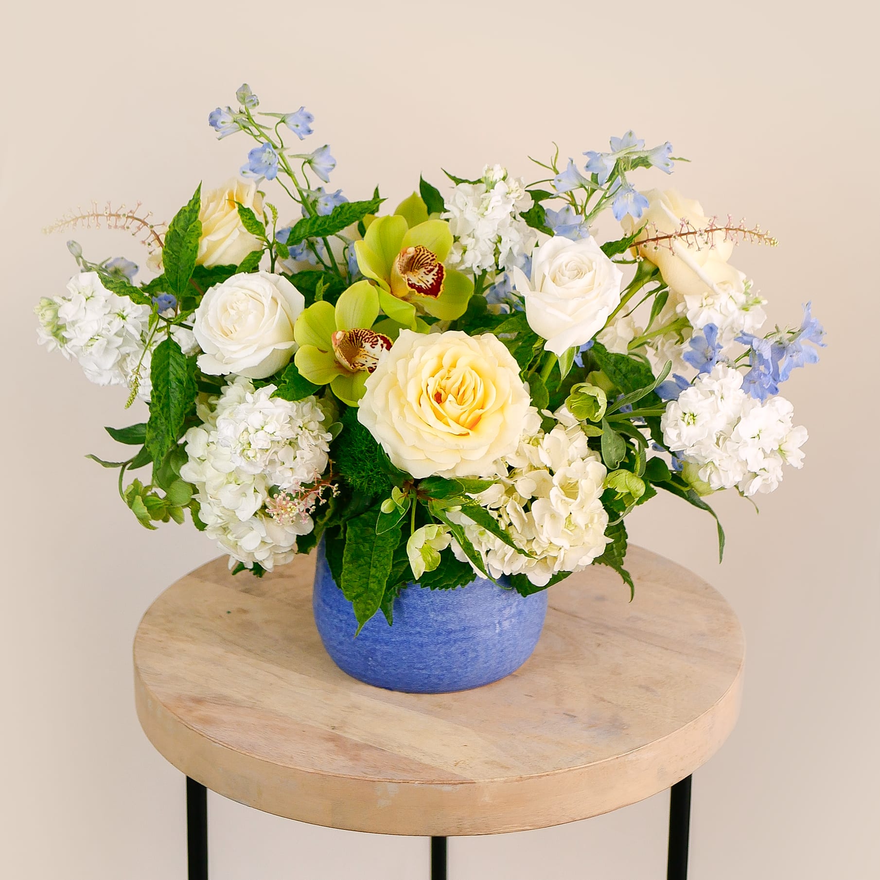 Santorini - A fresh and cheerful arrangement inspired by the colors of the Mediterranean. &quot;Santorini&quot; features cream and white roses, green cymbidium orchids, hellebores, light blue delphinium, white hydrangeas and more. Housed in a cerulean ceramic vase that evokes the iconic blues of Santorini’s rooftops, this design is perfect for brightening up any room! Arrangement approximately measures 12&quot; tall and 15&quot; wide with flowers.