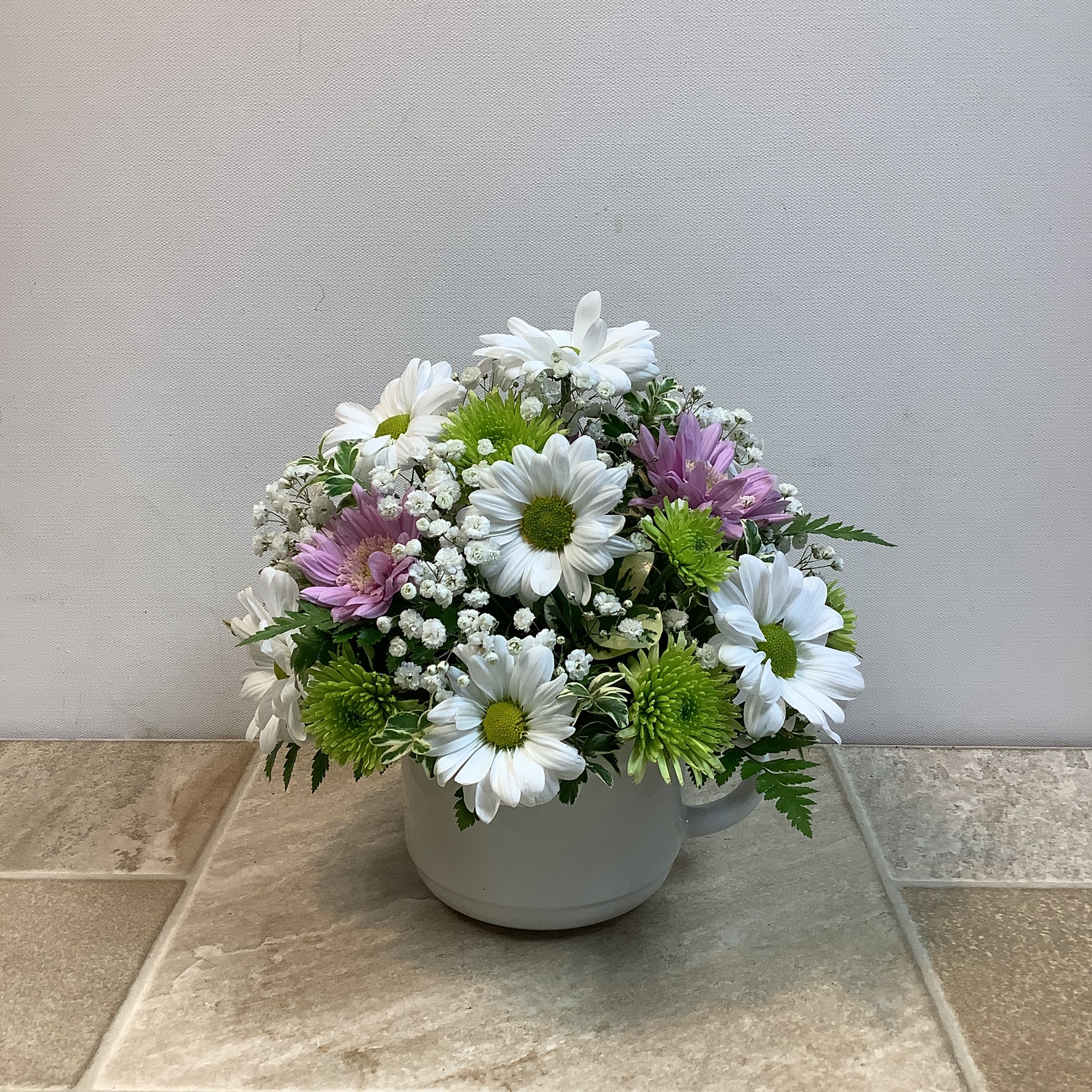 Sweet and Petite - This petite bouquet is a sweet and cheerful floral arrangement displayed in a simple blue container. It features a charming mix of blooms including soft lavender daisy mums, crisp white daisies, and bright green athos.  The flowers are accented with delicate sprigs of baby’s breath and lush fern greenery, creating a fresh and airy look.  Compact in size and beautifully balanced, this bouquet radiates a light, uplifting feel — perfect for brightening up a desk, bedside table, or as a thoughtful “just because” gift. It measures 10” wide x 8” high.   