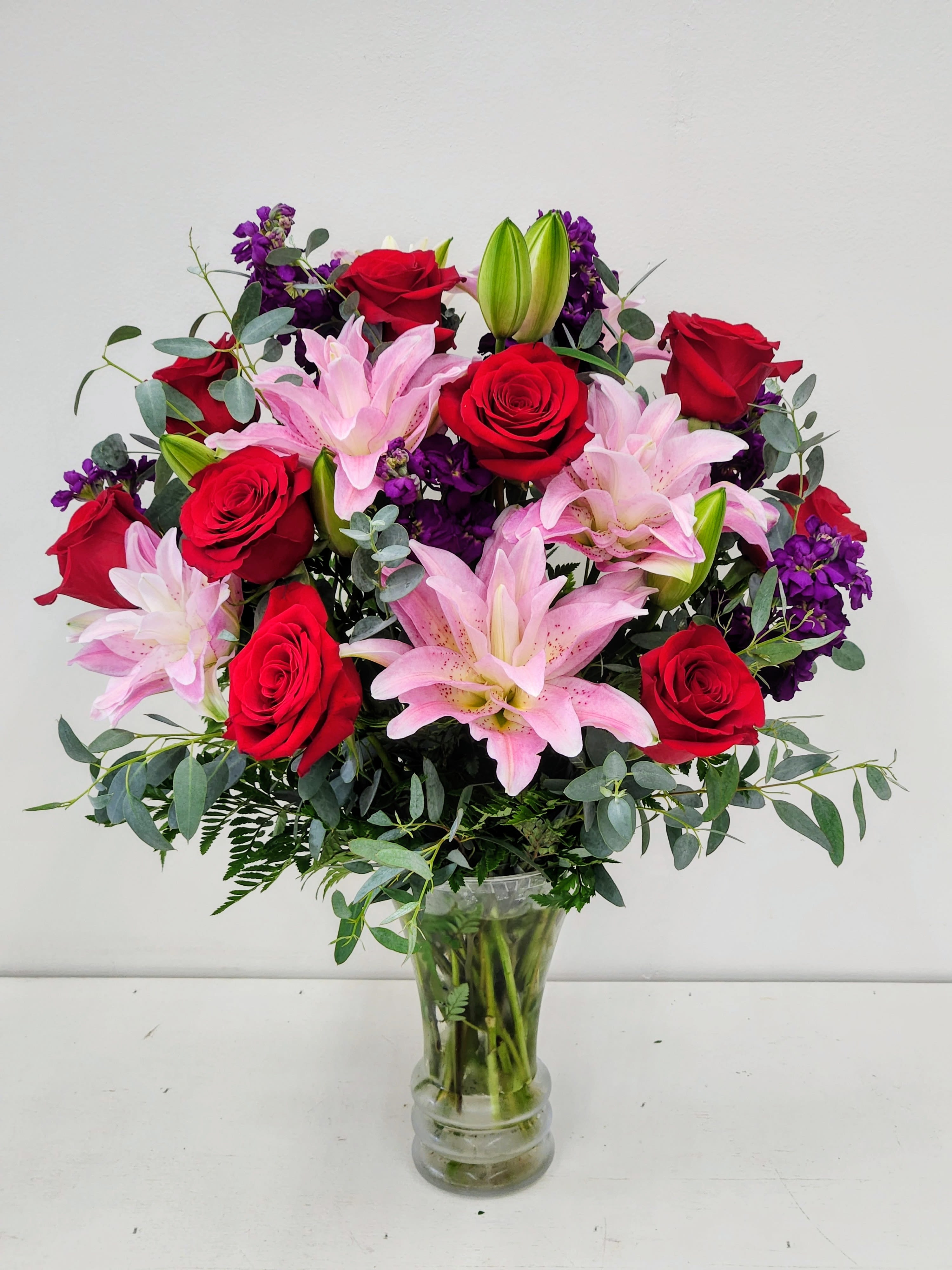 Blushing Beauty - Blush meets bold in this enchanting arrangement! Velvety red roses steal the spotlight alongside delicate pink rose lilies and charming purple stocks, all nestled in a sleek glass vase. Wispy eucalyptus leaves add an airy, graceful touch that softens the whole composition. Perfect for celebrating love, brightening someone's day, or simply treating yourself to a moment of floral joy.