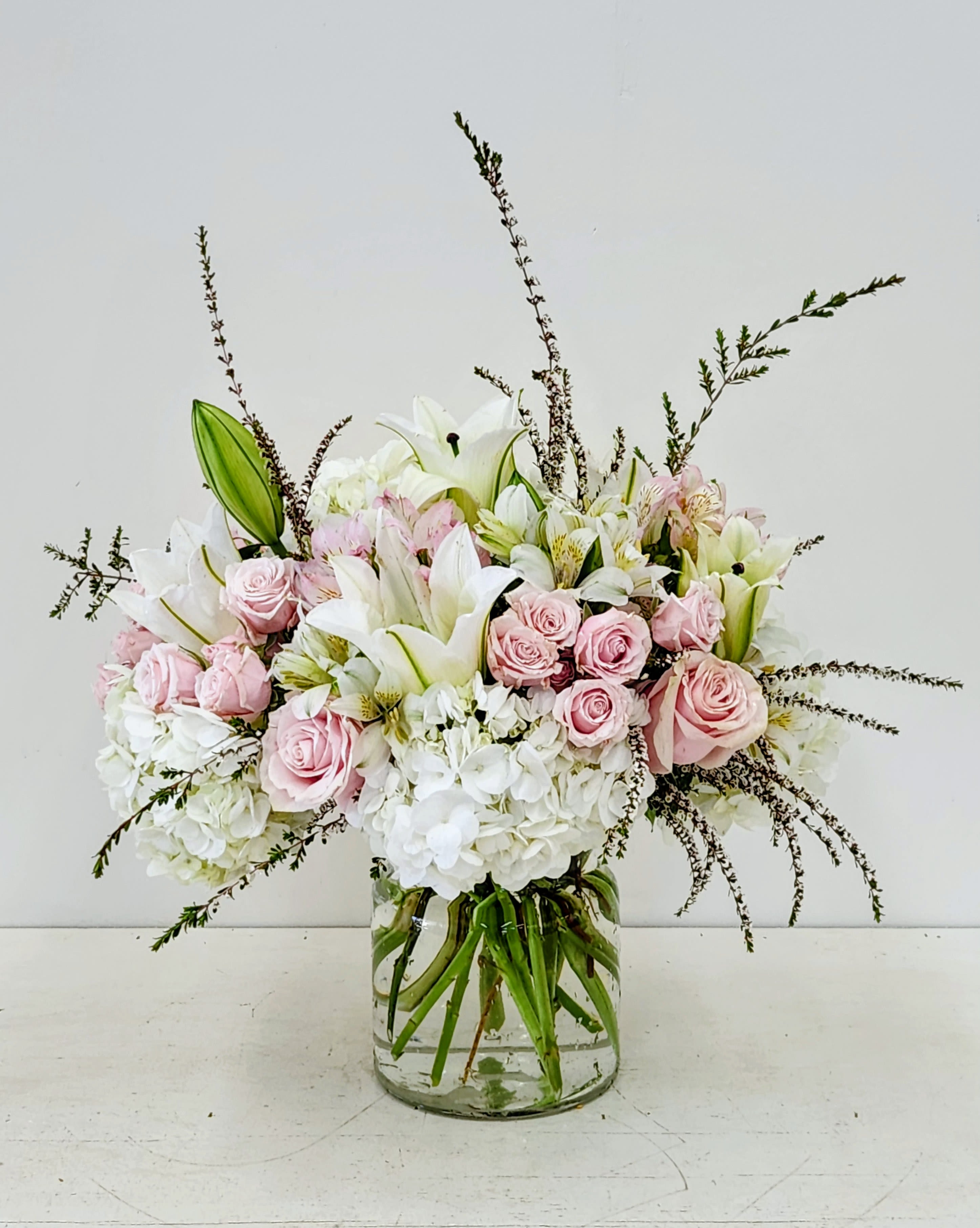 Simple Elegance - Discover understated charm with our Simple Elegance arrangement. Pristine white hydrangea forms a lush foundation, complemented by delicate pink spray roses that add a whisper of warmth. Dainty white lilies bring refinement, while seasonal wisps and tall textural elements create dimension and movement. Perfect for those who appreciate beauty in simplicity—an arrangement that speaks volumes through graceful restraint.  AS PICTURED: PREMIUM 15&quot; WIDE by 22&quot; TALL