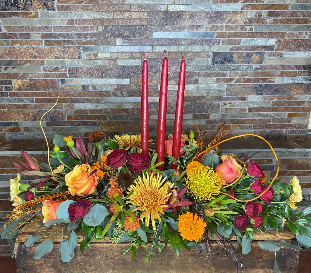Harvest Blessings - Our largest centerpiece option that is perfect for big family gatherings and long dining tables. Three burgundy candles set amidst traditional colors designed long and low. Premium flowers will be hand selected by our designers to give the arrangement it's perfect flair. Take your dinner table to the next level this holiday season.  