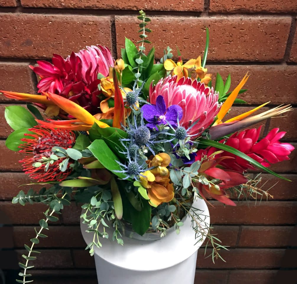 Wildly Tropical Bowl - Escape to the islands with this tropical display of flowers, featuring vibrant Proteas, Birds of Paradise, Orchids, Ginger and other fine imports. Hand-arranged with fresh garden greens in a bubble bowl vase. Standard size is approximately 16in (W) x 15in (H). Deluxe version is larger and features more premium blooms arranged in a larger bubble bowl vase.  • Standard - 2 Red Gingers, 2 Pink Ice Proteas, 1 Pincushion Protea, 3 Safari Sunset Proteas, 3 Birds of Paradise, 3 Mokara Orchids, 2 Blue Thistle, Eucalyptus and Fresh Garden Greens - 8in Bubble Bowl Vase  • Deluxe - 3 Red Gingers, 3 Pink Ice Proteas, 2 Pincushion Protea, 4 Safari Sunset Proteas, 4 Birds of Paradise, 3 Mokara Orchids, 3 Blue Thistle, Eucalyptus and Fresh Garden Greens - 10in Bubble Bowl Vase  Care Tips: Place your bouquet in a cool location away from direct sunlight, heating or cooling vents, drafty places, directly under ceiling fans, or on top of televisions or radiators. Check water level daily, keep the vase full with clean water. Change water every 2-3 days and apply a sharp fresh cut to the stems to ensure extended flower's life span.