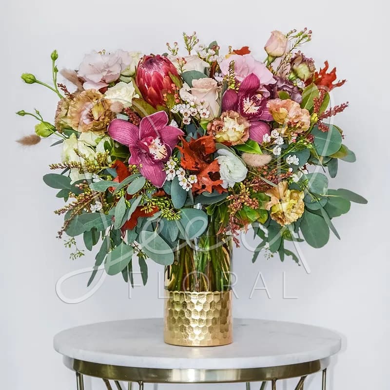 Seasonal Autumn Arrangement - Celebrate the beauty of fall with our Seasonal Autumn Arrangement, a stunning blend of warm, earthy tones and lush seasonal blooms. Featuring elegant orchids, protea, garden roses, lisianthus, and fall foliage, this tall floral design captures the cozy, sophisticated essence of autumn. Arranged in a textured gold vase, it adds a touch of luxury to any dining table, entryway, or event.  Perfect for Thanksgiving centerpieces, fall home décor, or seasonal gifting, this arrangement brings the richness of autumn indoors. 