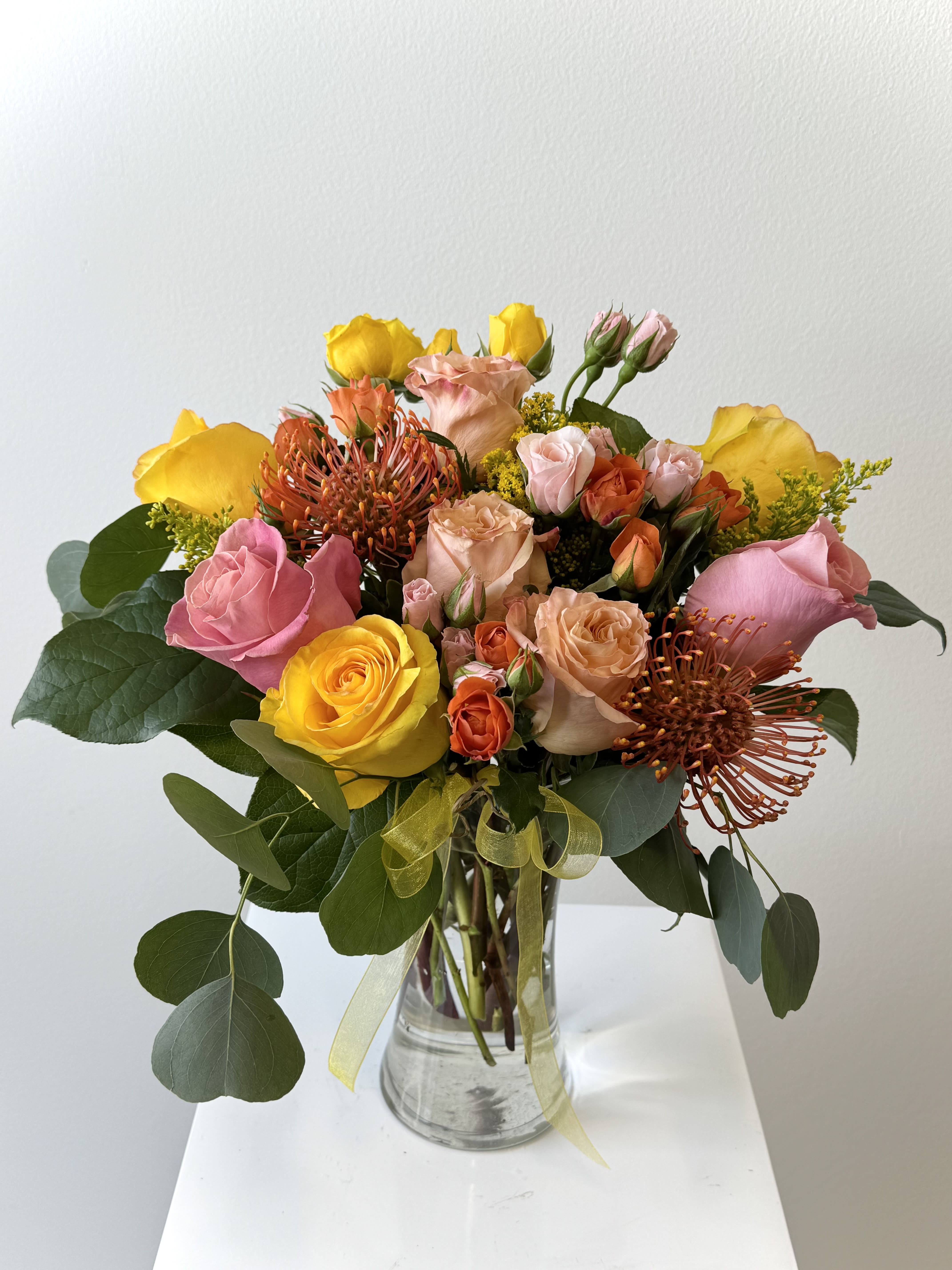 Sunburst - Sunburst Flower Arrangement – Vibrant Pincushion &amp; Multicolor Rose Bouquet with Solidago Ignite joy with the “Sunburst” flower arrangement, a dazzling explosion of summer hues! Bold pincushion protea takes center stage with its spiky, firework-like texture, surrounded by radiant yellow, pink, and orange roses in full bloom. Golden solidago weaves through like rays of sunlight, creating a compact, high-impact design that screams celebration and energy.  Why Sunburst Shines: Pincushion Protea: Exotic, long-lasting focal flower symbolizing courage and transformation. Yellow Roses: Burst with friendship, joy, and new beginnings. Pink Roses: Convey gratitude, appreciation, and sweet admiration. Orange Roses: Ignite passion, enthusiasm, and pride. Solidago: Fluffy golden filler adds warmth, volume, and natural glow.  Hand-arranged in a modern glass vase. Perfect for birthdays, congratulations, promotions, housewarmings, or “you’ve got this” encouragement. Sunburst flower arrangement, pincushion protea bouquet, yellow pink orange roses, solidago floral design, colorful rose delivery, protea rose bouquet, birthday sunburst flowers, congratulations floral gift, same-day vibrant bouquet, modern flower arrangement. Order “Sunburst” now and deliver instant happiness! Same-day delivery available. Sunburst bouquet, pincushion protea, multicolor roses, solidago flowers, vibrant florals.