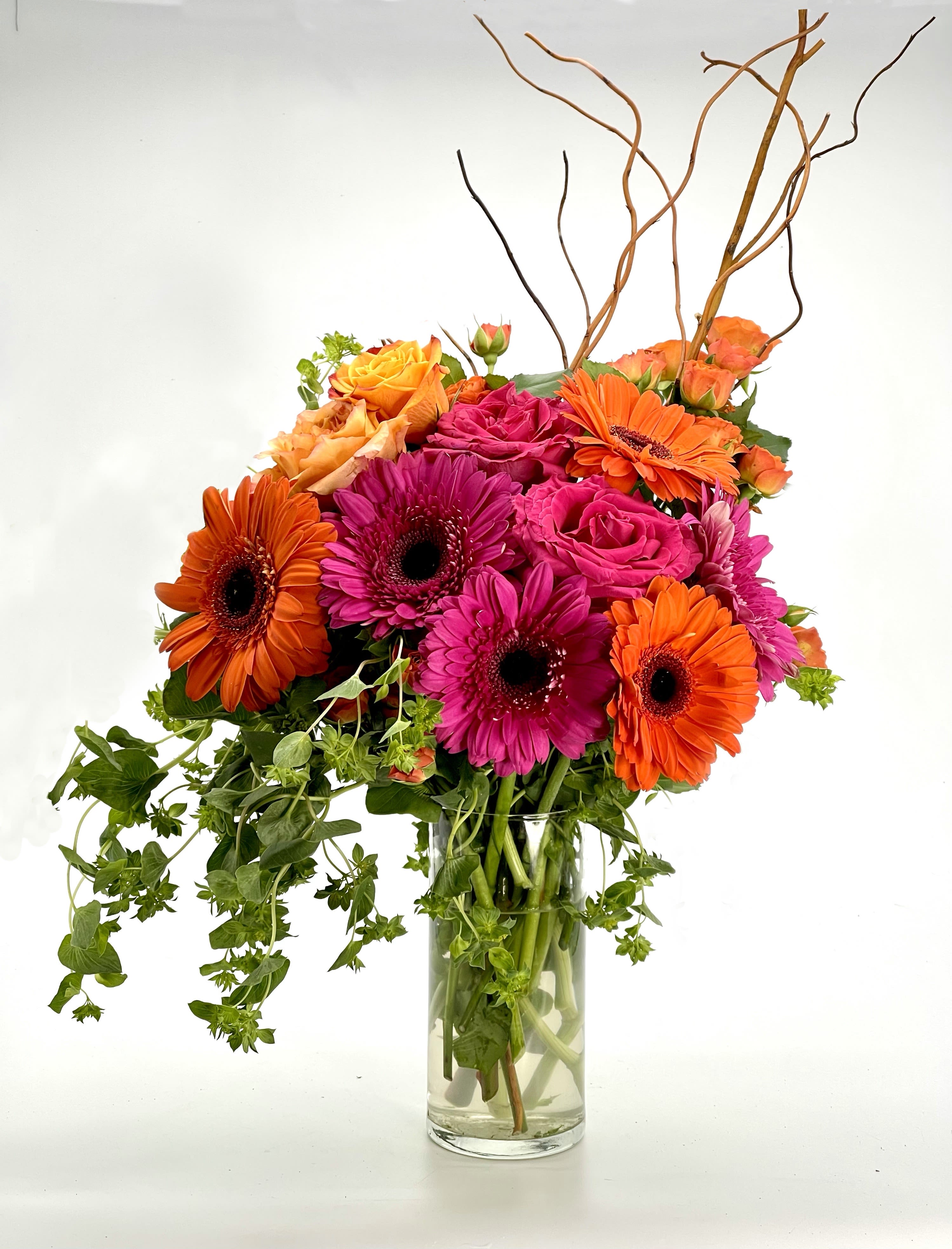 Spring Breeze Bouquet - A vibrant floral arrangement featuring bold orange gerbera daisies, bright pink roses, and lush greenery in a sleek glass vase. Perfect for adding a burst of color and elegance to any space or occasion.