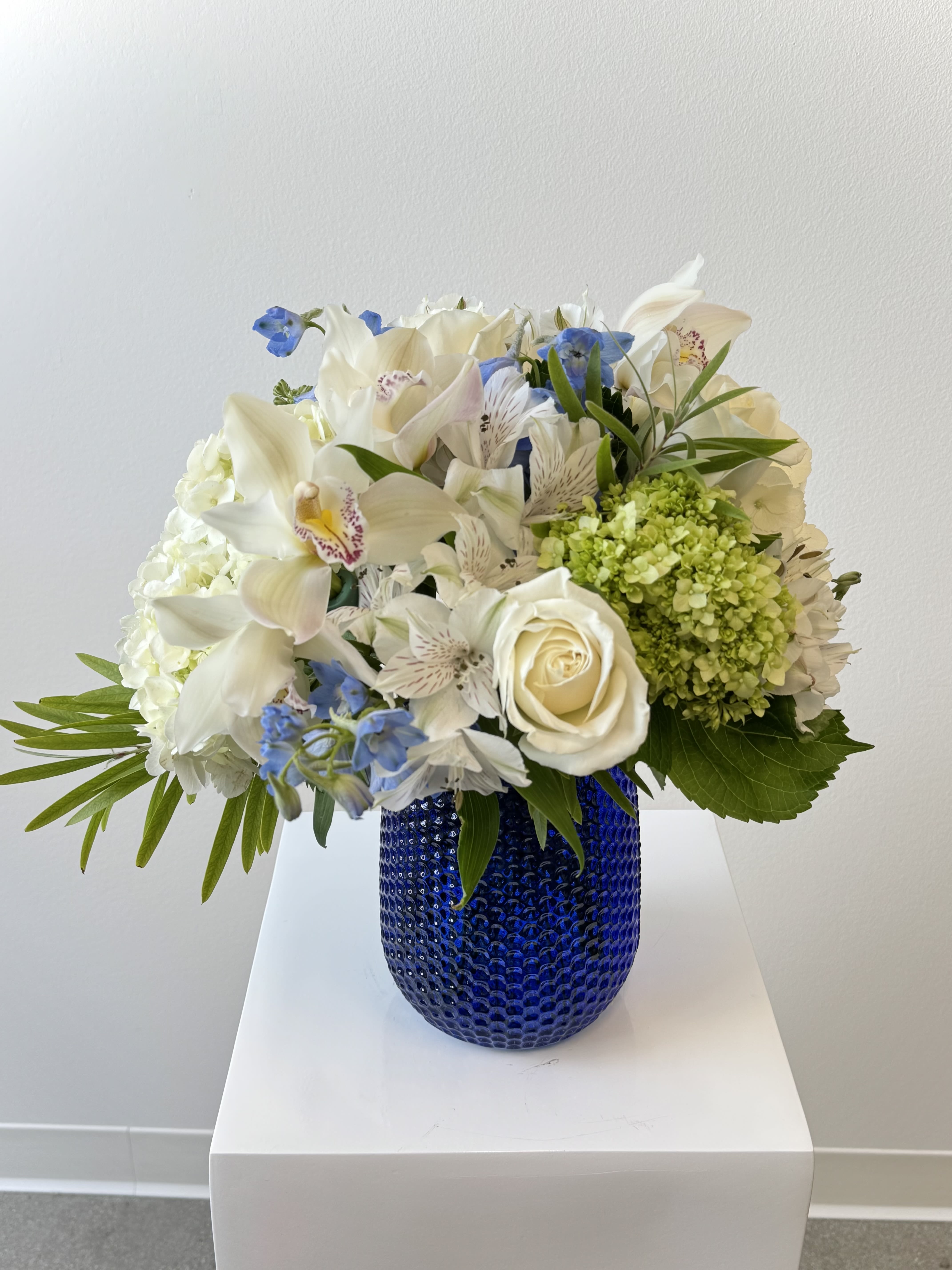 Royal in Blue - Royal in Blue Flower Arrangement – An Elegant Blue Delphinium &amp; White Orchid Bouquet Step into regal serenity with Royal in Blue, a majestic symphony of cool whites and striking sapphire blues. Towering delphinium spires command attention, framed by voluminous white &amp; green hydrangeas, exotic white cymbidium orchids, delicate white alstroemeria clusters, and timeless white roses. This sophisticated arrangement evokes royal gardens and effortless luxury.  Why Royal in Blue Reigns Supreme: Delphinium: Dramatic blue spikes symbolize dignity and grace. White &amp; Green Hydrangeas: Lush, cloud-like blooms for serene volume. White Cymbidium Orchids: Premium, long-lasting elegance with tropical allure. White Alstroemeria: Airy filler extends freshness and texture. White Roses: Classic symbols of purity, unity, and new beginnings.  Hand-arranged in a striking blue vase. Perfect for weddings, corporate events, sympathy tributes, anniversaries, or luxury home décor. Royal in blue flowers, blue delphinium bouquet, white hydrangea orchid arrangement, cymbidium alstroemeria roses, luxury white blue floral, wedding delphinium centerpiece, sympathy elegant bouquet, corporate blue flowers, same-day royal flower delivery, tall blue vase arrangement. Crown any occasion with Royal in Blue! Same day delivery available. Royal In Blue, delphinium bouquet, white orchids, luxury floral, blue white wedding.