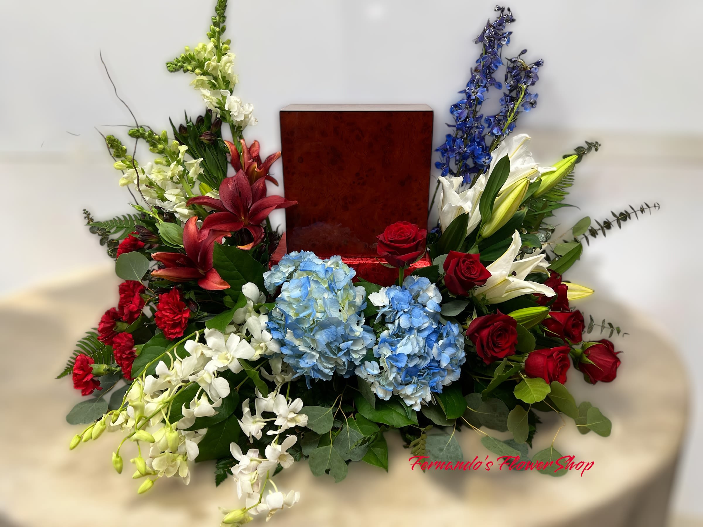 Always in my heart  - The image showcases a floral arrangement designed around a wooden urn, making it a memorial or funeral display. The arrangement is composed of various flowers, including red roses, red lilies, blue hydrangeas, white lilies, white snapdragons, white orchids, and red carnations. There are also some blue delphiniums adding contrast to the design.  The flowers are artistically arranged with a mix of colors—deep reds, bright whites, and soft blues—symbolizing love, purity, and remembrance. The greenery, including eucalyptus leaves and other foliage, adds texture and depth to the composition. The urn, placed centrally, is surrounded by the flowers, making it the focal point of the arrangement.  The text “Fernando’s Flower Shop” is subtly overlaid on the bottom right corner of the image, indicating the florist responsible for this arrangement. The setting appears to be a neutral background, with the arrangement placed on a round, draped table.
