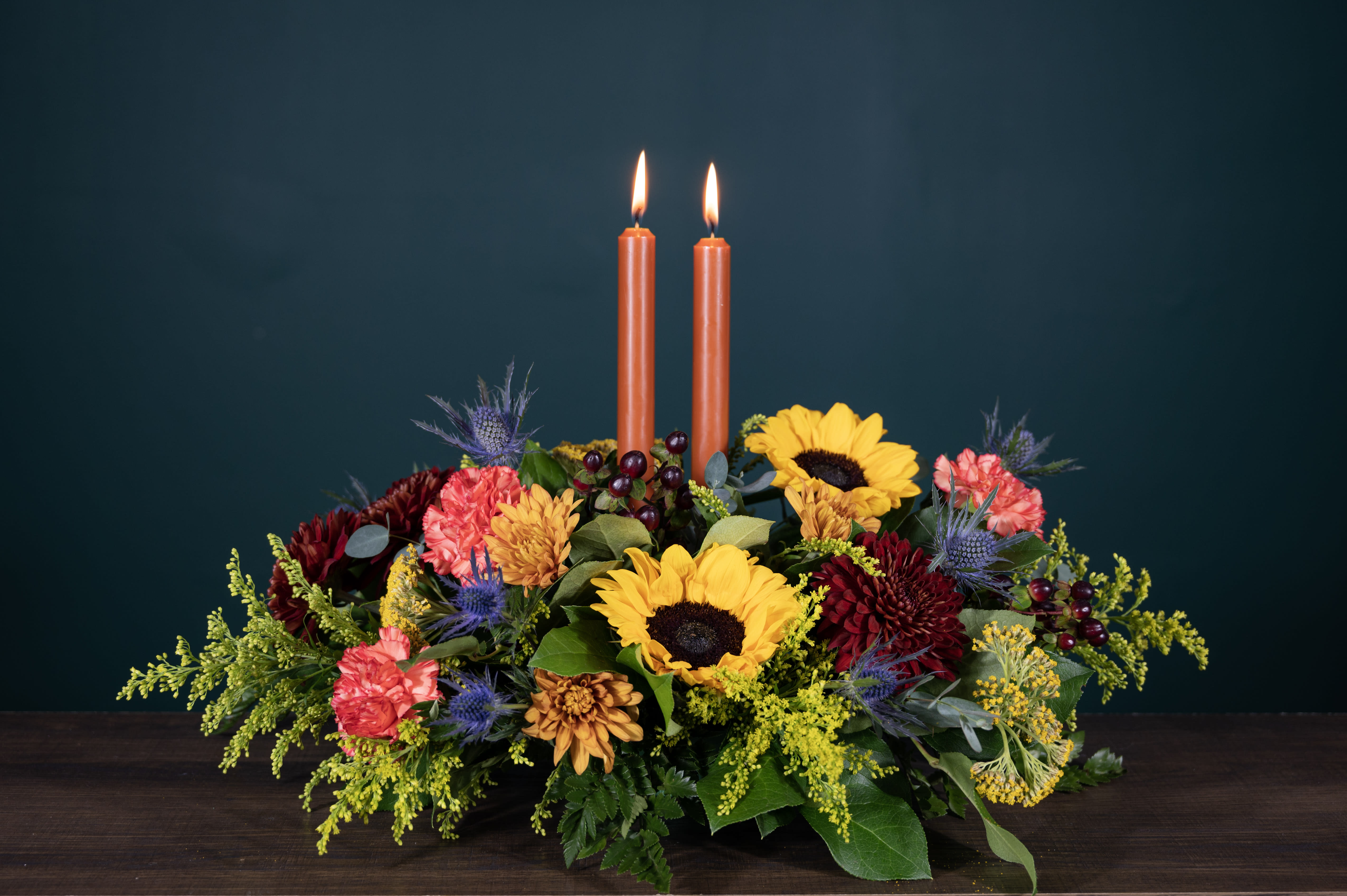 Grace Thanksgiving Centerpiece - Beautify any table this Thanksgiving season with the Grace Thanksgiving Centerpiece. Deep orange candles mixed with a beautiful array of sunflowers, mums, berries and thistle. If you can't be there in person, send this reminder of your love.   Product Details: This arrangement is approximately 15&quot; long and 6&quot; tall (candles extend 15&quot; tall.) Candles are dripless. 