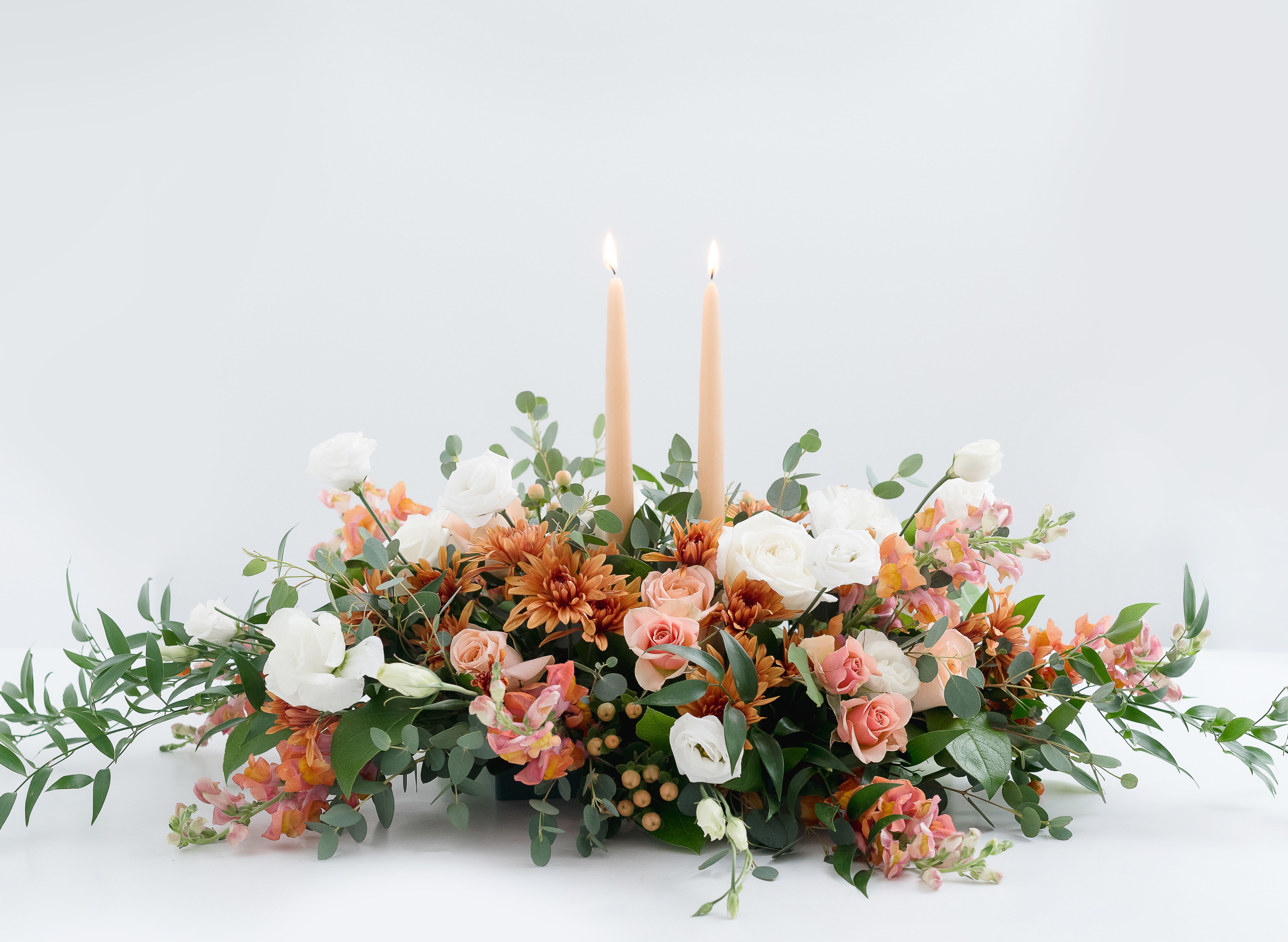 White Pumpkin Thanksgiving Centerpiece  - A stunning design for any holiday table. An elegant and soft take on Fall colors for the sophisticated table. Roses, mums, Snapdragons and berries with Eucalyptus make up this centerpiece with Gold Spice taper candles. The perfect gift to send or to adorn your Thanksgiving table.   Product Details: This arrangements is approximately 18&quot; long and 10&quot; wide. 