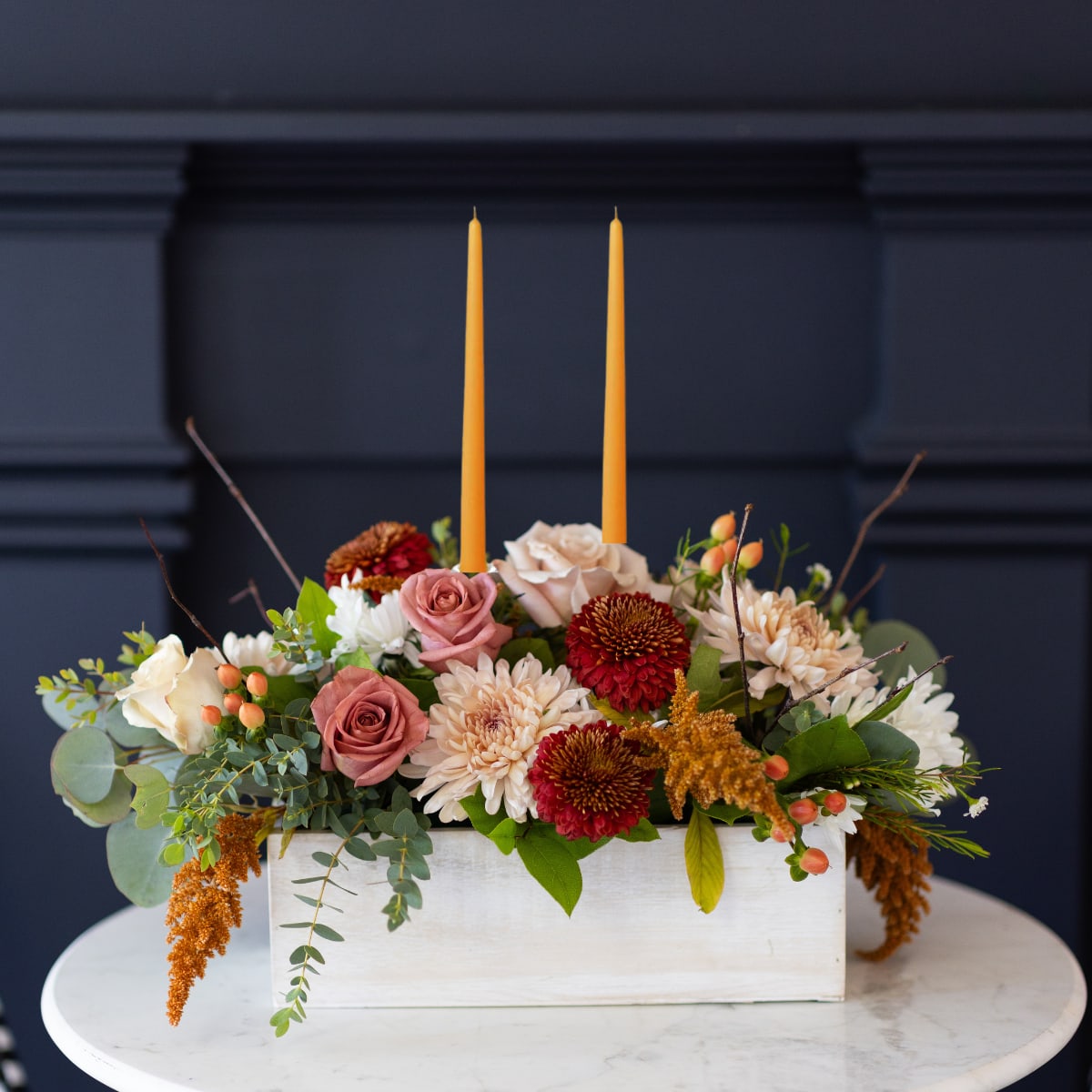 Chai Latte Centerpiece with Candles  - A lovely Thanksgiving centerpiece inspired by the cozy vibes of warm fall spices in shades of cinnamon, mocha, and cream in a seasonal mix of mums, roses, berries, and branches—all beautifully arranged in a white-washed wood box with matching candles.   Perfect as a gift for a friend or loved one, or as a charming centerpiece for a fall feast.  Product Details: This arrangement is 14&quot; long and 7&quot; wide with dripless candles. The DeLuxe size includes an additional 7 stems of flowers and greenery. 