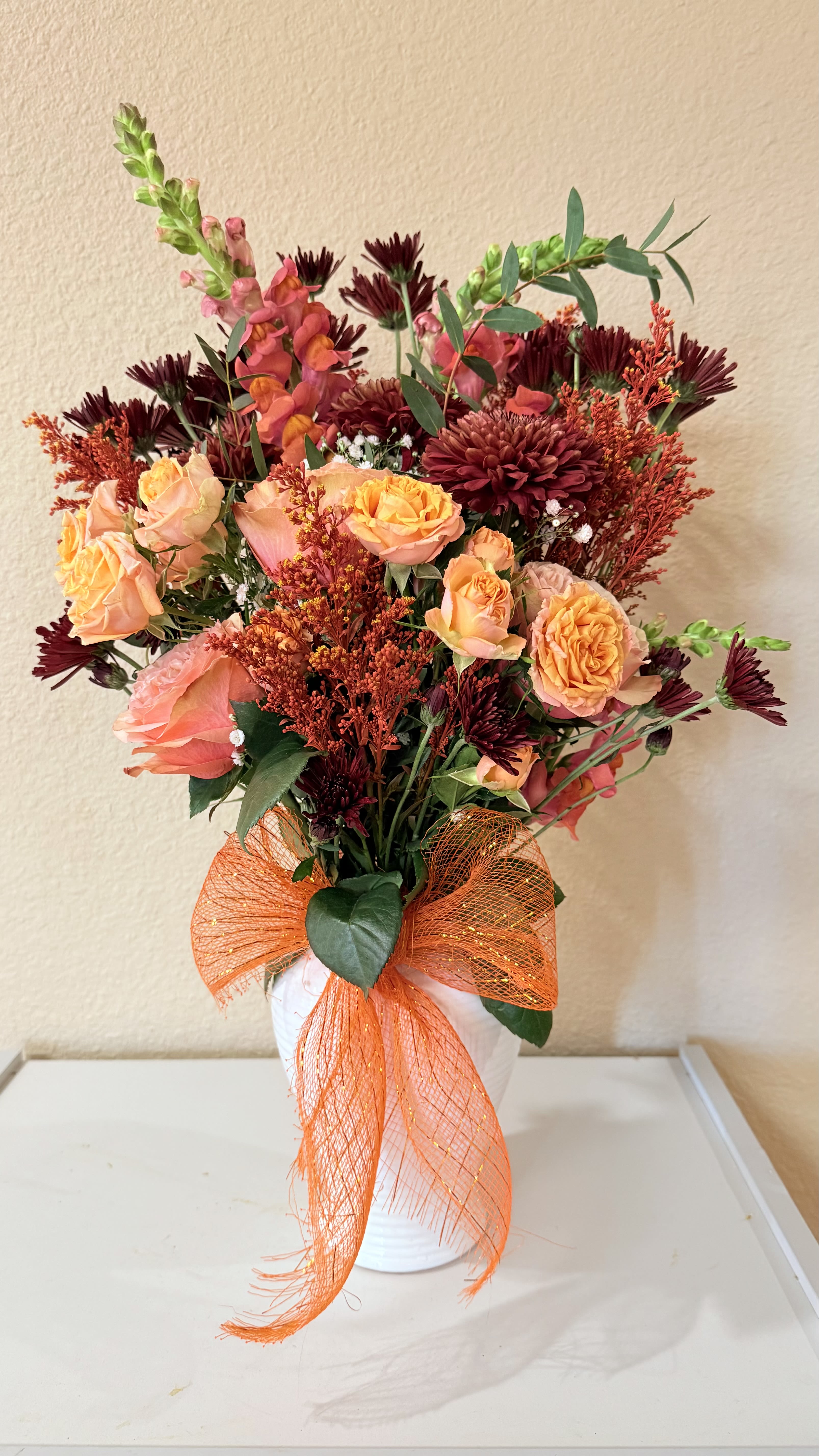 Grateful  Glow  - Rich burgundy blooms meet soft peach roses, accented with snapdragons, greenery, and a festive bow. A stunning choice for Thanksgiving, celebrations, or simply bringing warmth into your home.