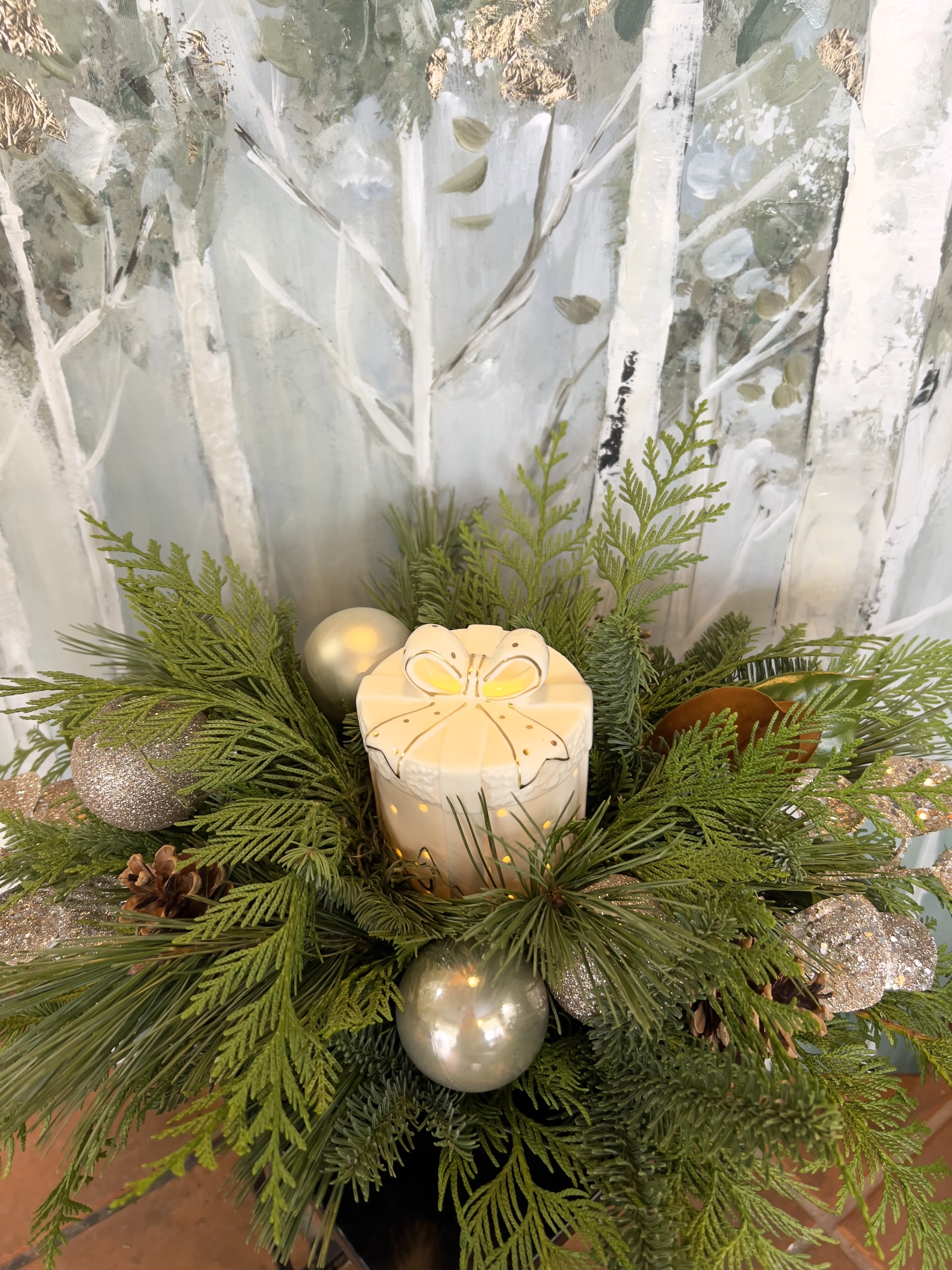 Twinkling Treasures - This arrangement incorporates a ceramic holiday present with a soft warm glow, includes glimmering holiday decorations, winter greens, and pine cones.