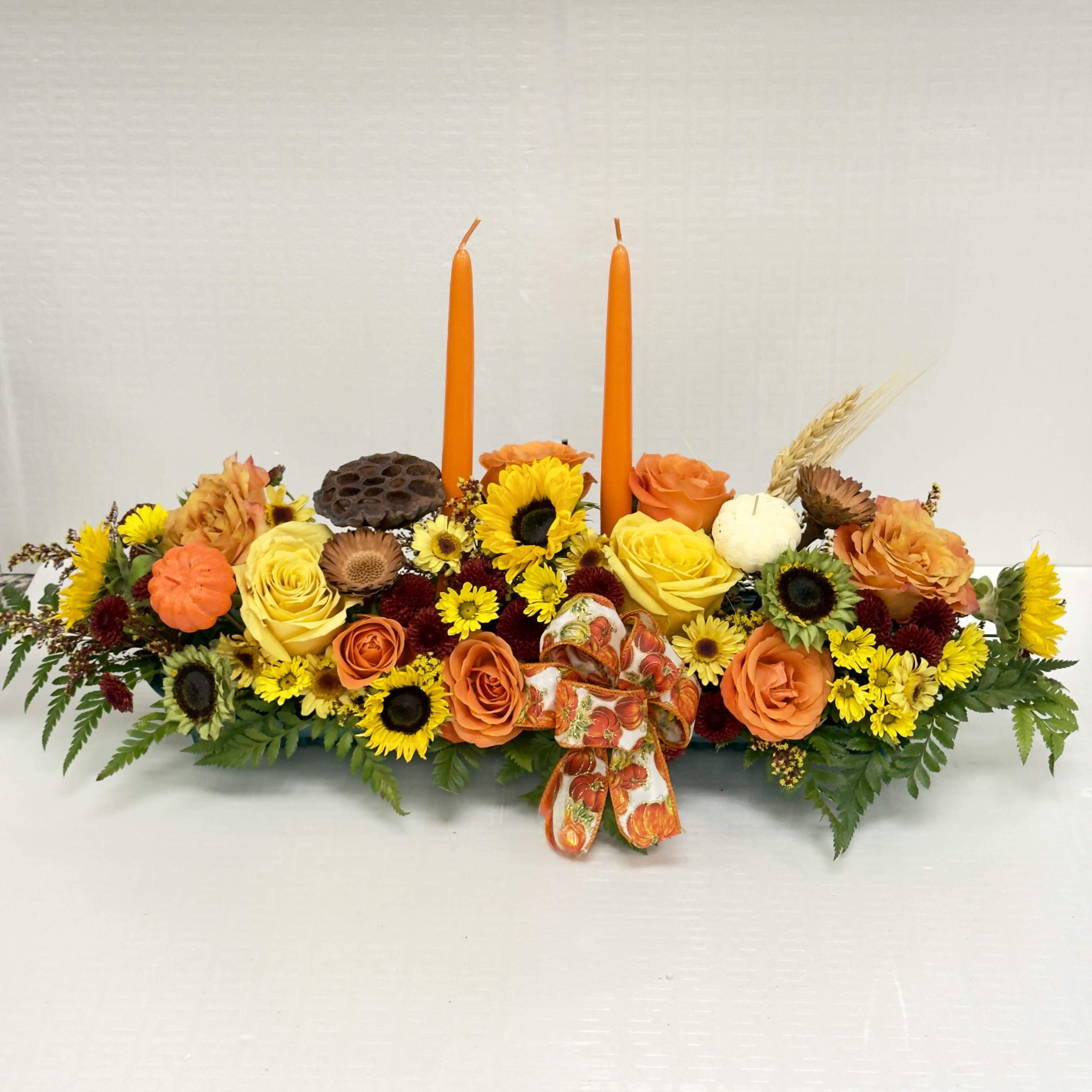 Harvest Candle Centerpiece - A warm and festive arrangement featuring vibrant sunflowers, roses, autumn blooms, and seasonal accents, complete with glowing taper candles. Perfect for enhancing your Thanksgiving table or adding cozy fall charm to any gathering.