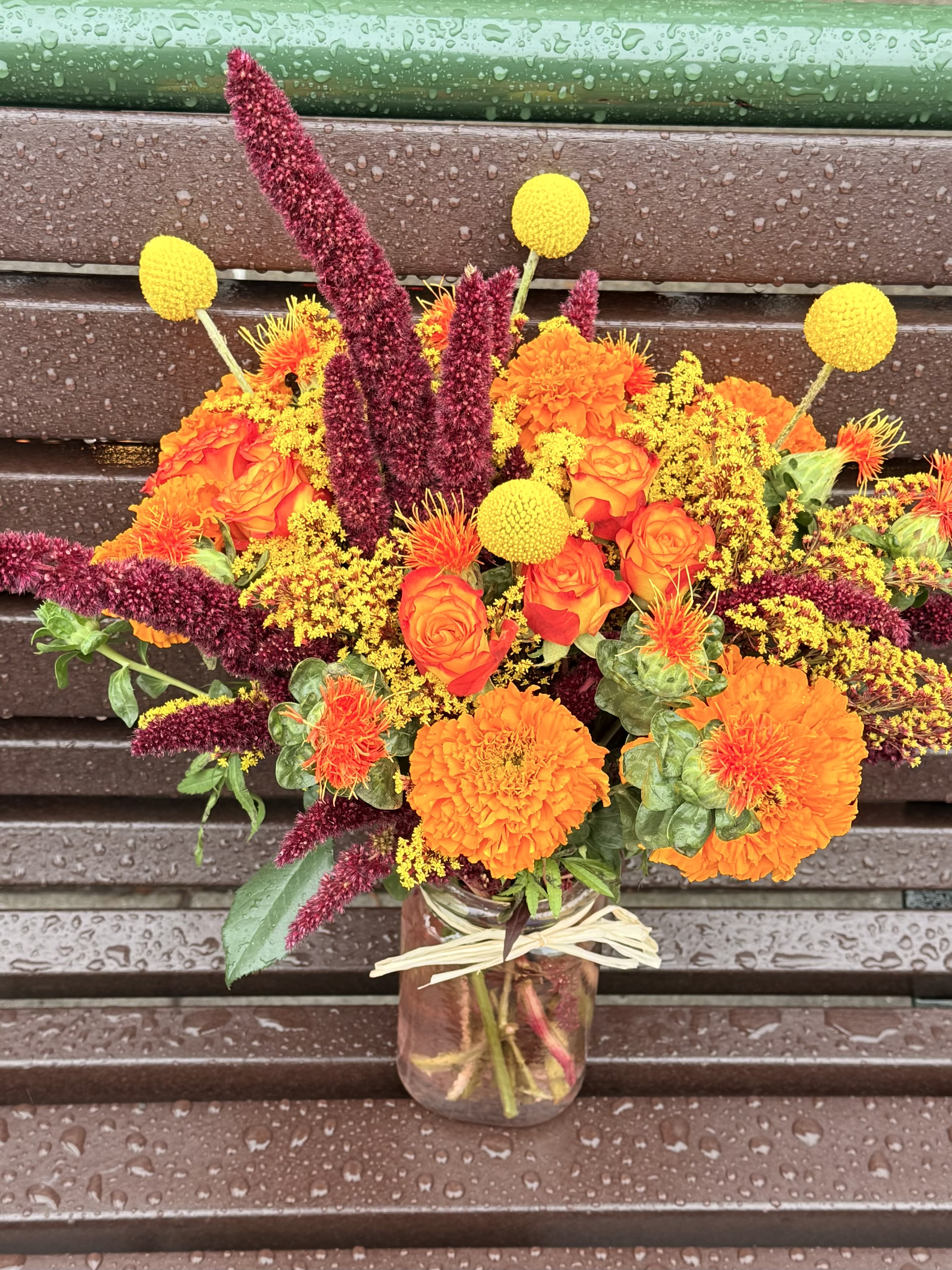 Autumn Ember  - A vibrant, fiery celebration of fall, Autumn Ember Harvest bursts with rich orange marigolds, safflower, golden craspedia, and sunset-toned spray roses. Deep burgundy amaranthus adds dramatic texture and height, while soft yellow solidago weaves warmth throughout the bouquet. Designed in a rustic mason jar with natural raffia, this arrangement captures the cozy glow of autumn and brings a touch of seasonal charm to any home or event.